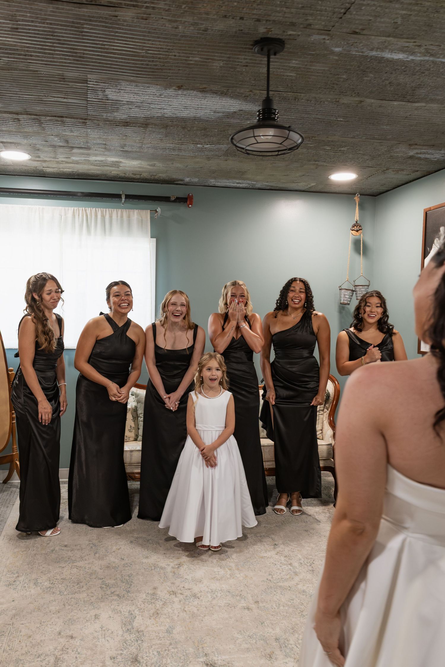 Bridesmaids in black dresses react emotionally seeing bride for first time with flower girl present.