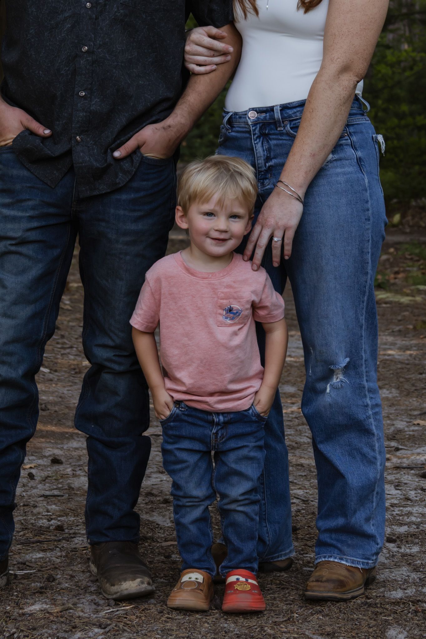 Young blonde toddler boy in pink shirt and jeans smiling between two adults in an outdoor family portrait.