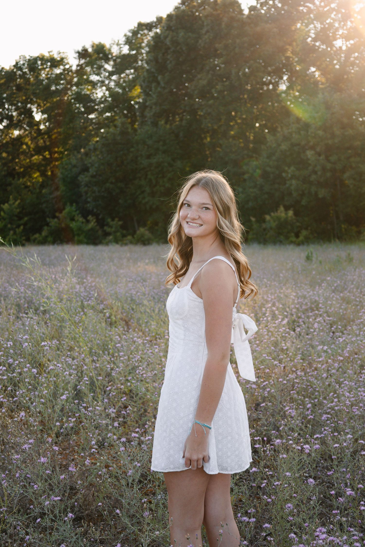 Young woman in white sundress smiling in golden hour sunlit meadow with wildflowers and trees.