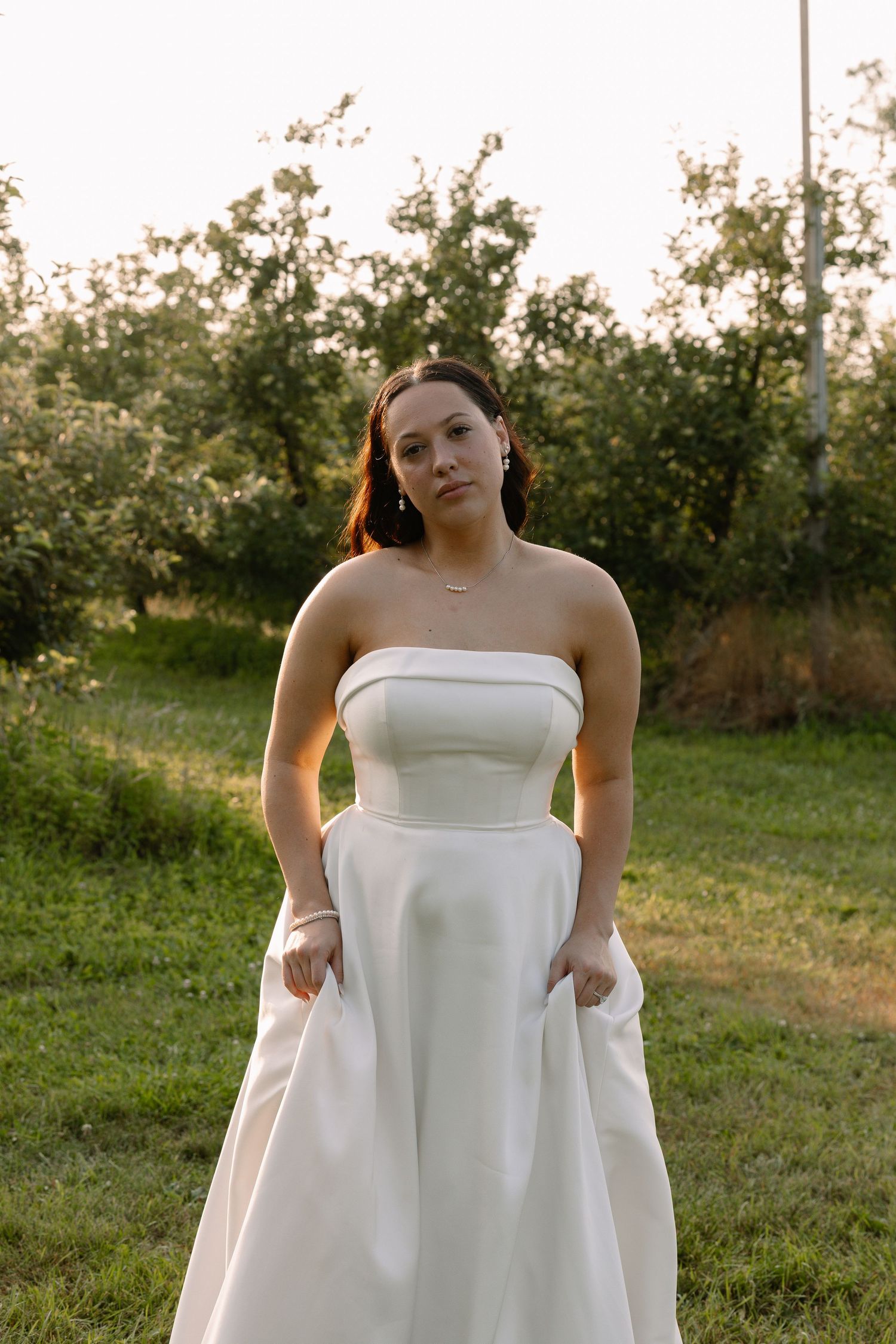 Bride in strapless white ball gown poses gracefully outdoors at golden hour in lush green orchard.