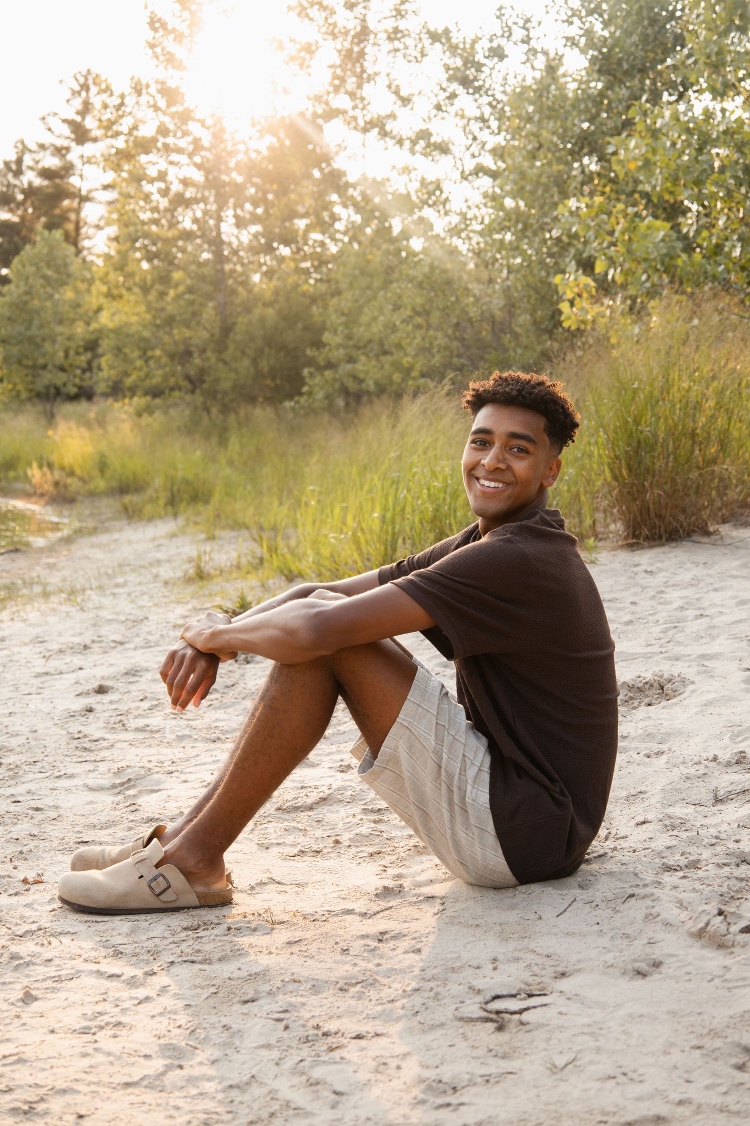 Smiling young man sitting on sandy ground outdoors, wearing dark shirt and shorts, with green trees in golden sunlight.