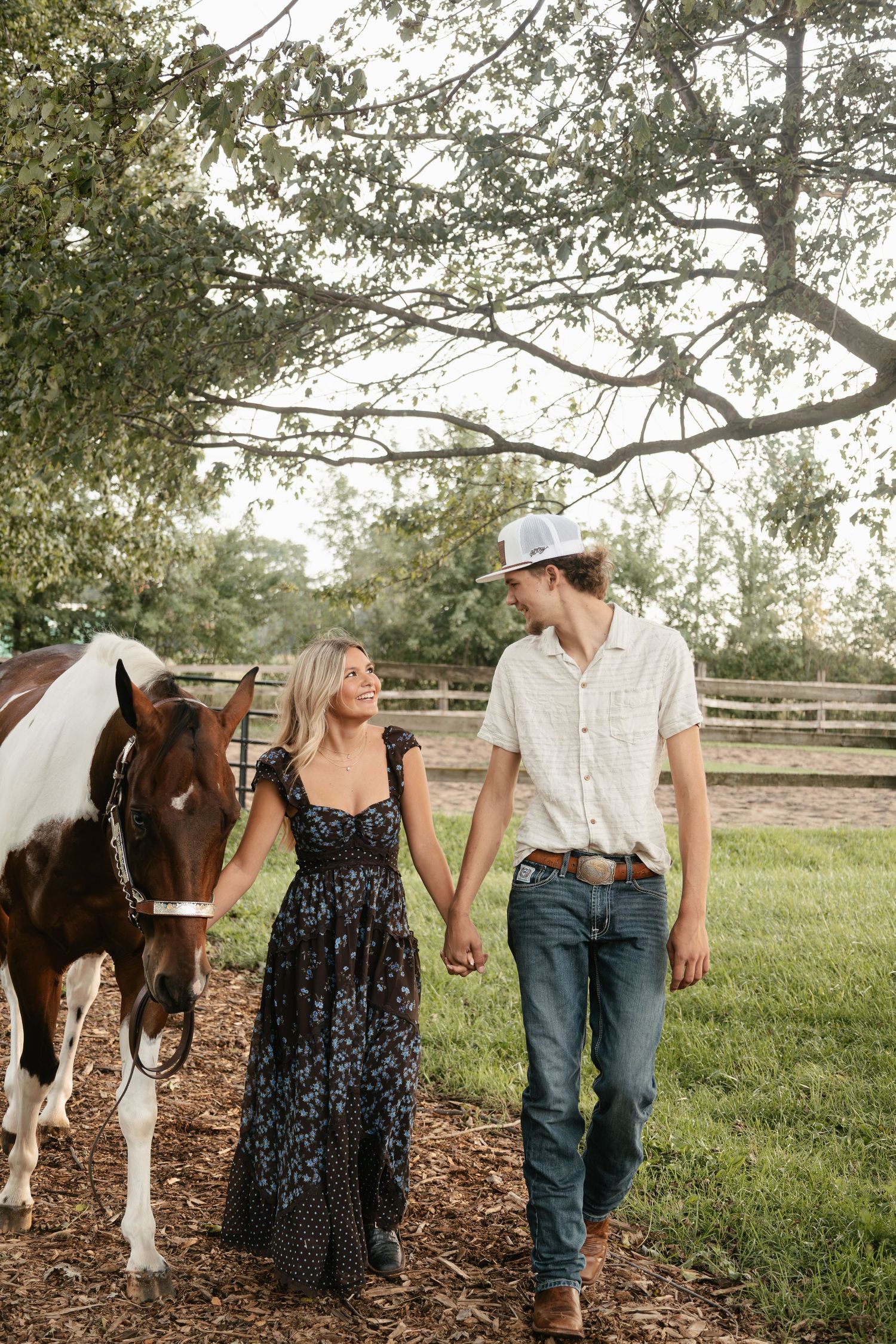 A couple holds hands and walks beside a paint horse on a farm, surrounded by lush green trees and a wooden fence.