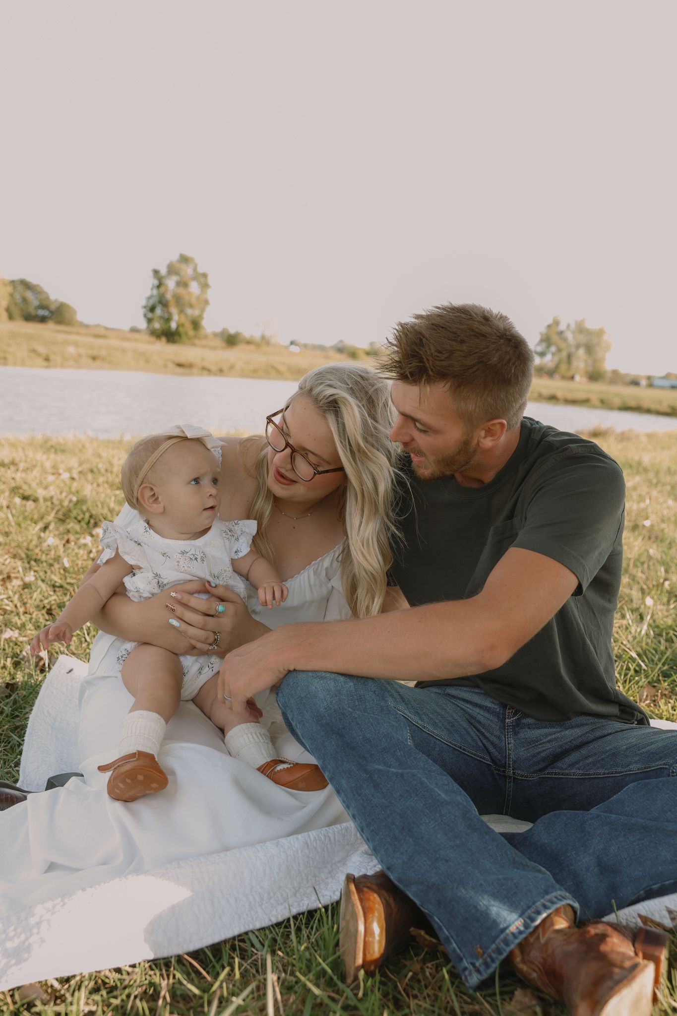 Happy couple sitting on white blanket in field gazing lovingly at their smiling baby girl outdoors.