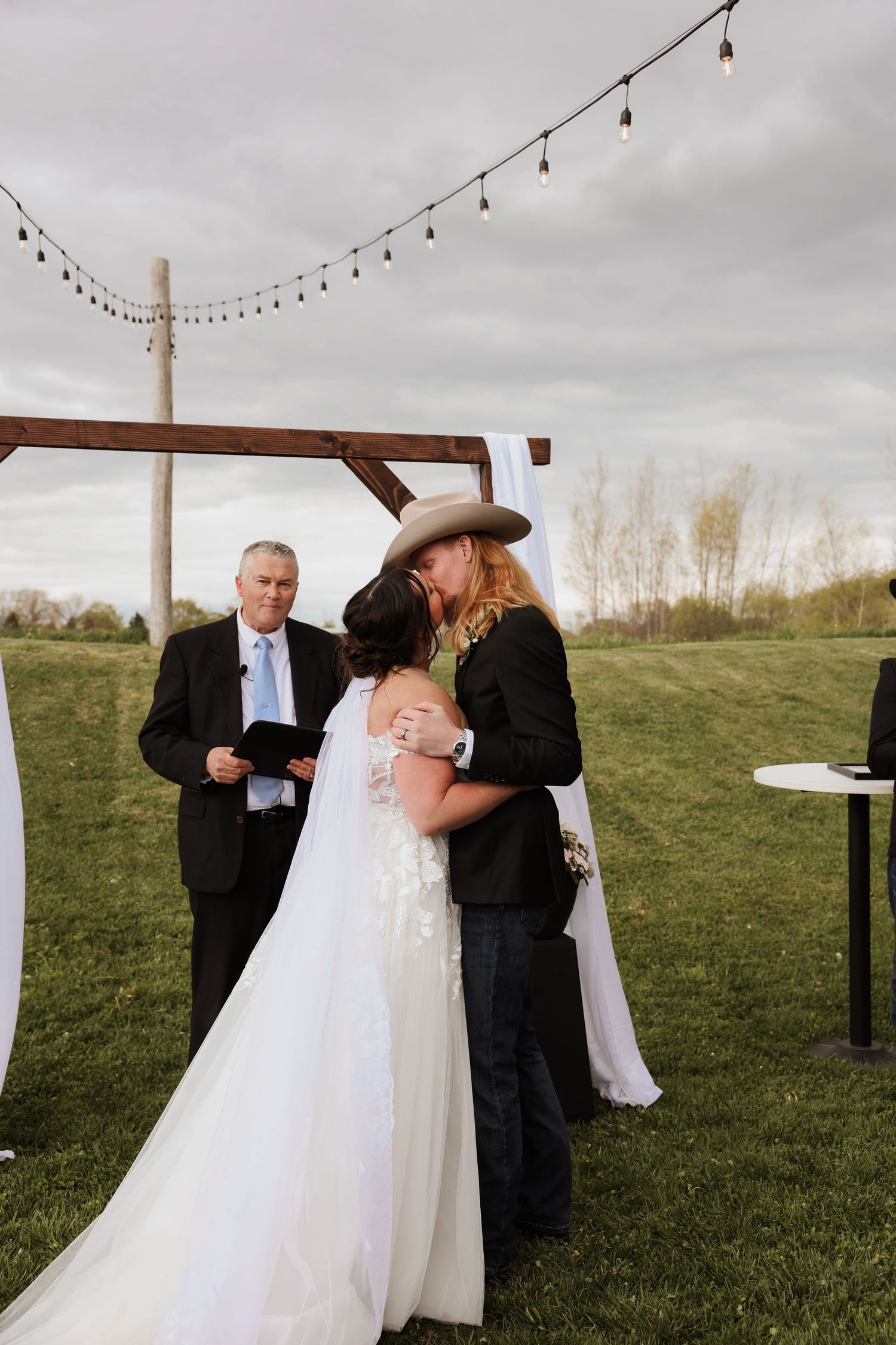 Bride and groom share first kiss at outdoor wedding ceremony under wooden arch with string lights.