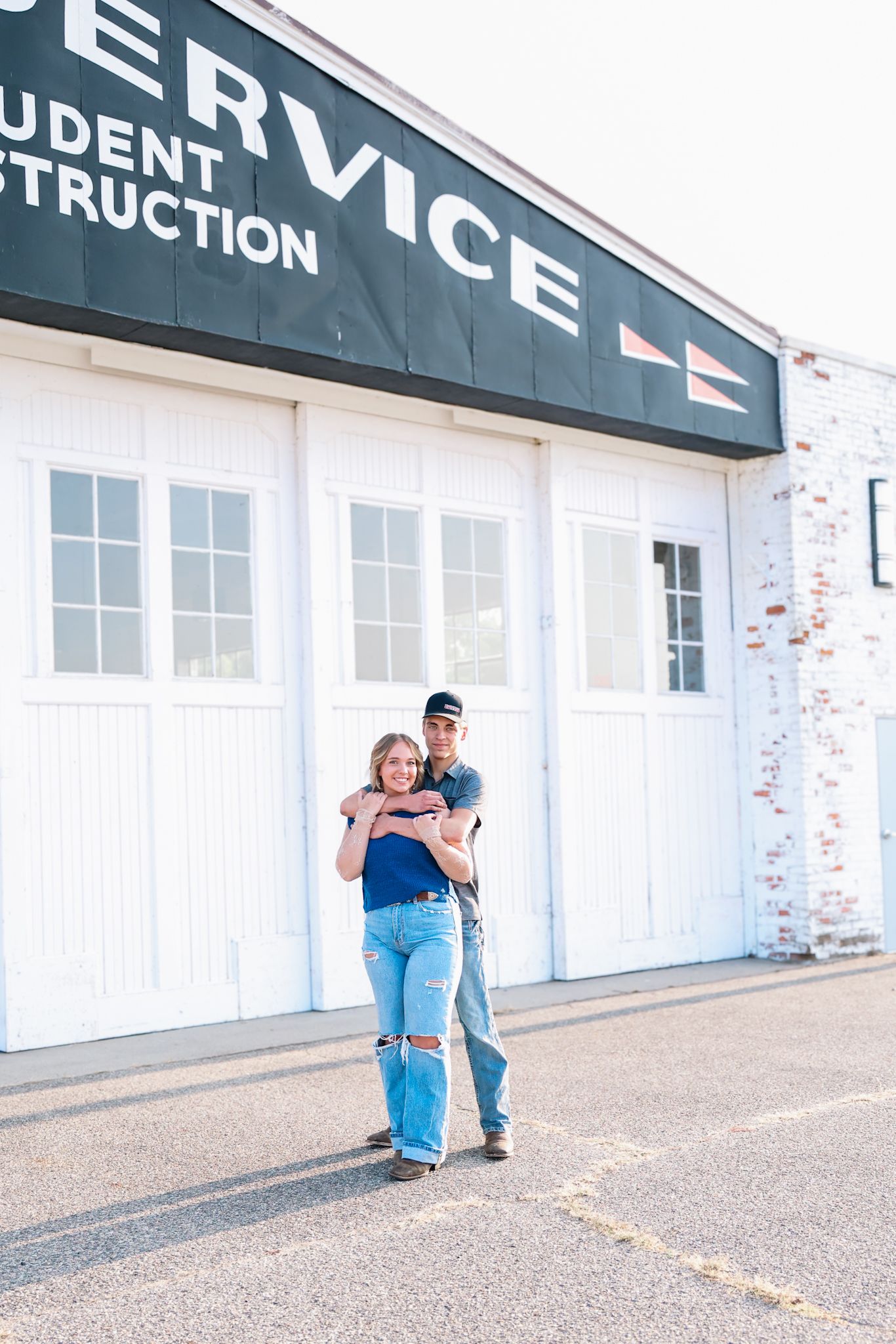 A couple poses together outside a Service building, the man embracing the woman from behind, both wearing jeans.
