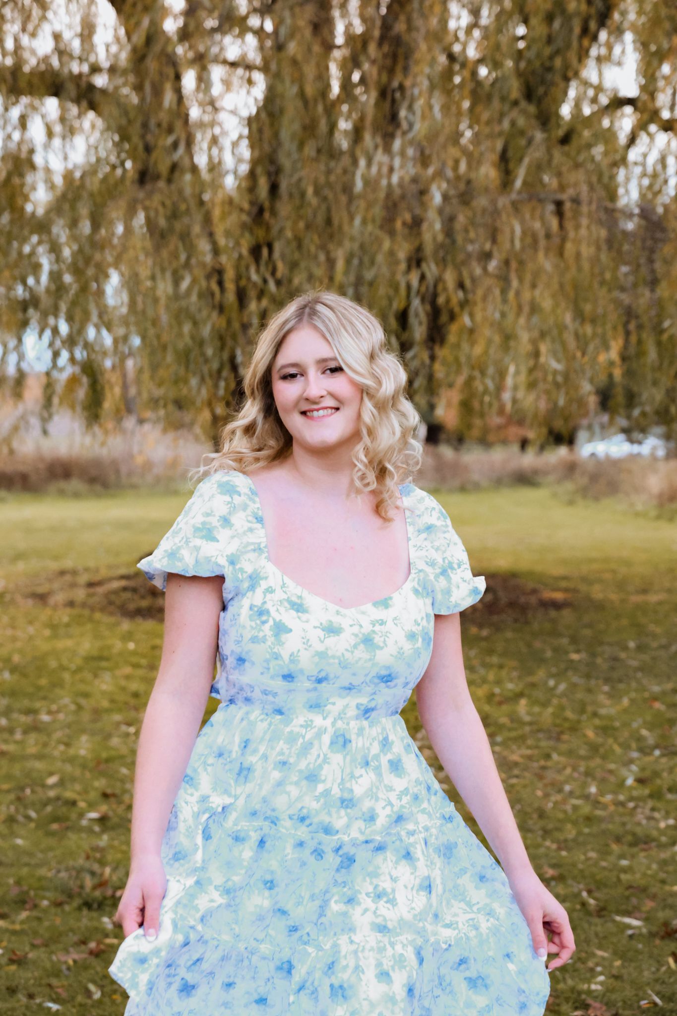 Smiling blonde woman in blue floral dress standing under weeping willow tree in park.