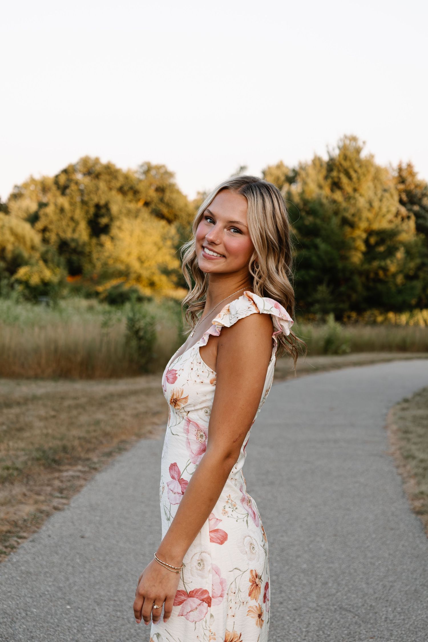 Young woman in floral dress smiling on a path surrounded by golden trees at sunset.