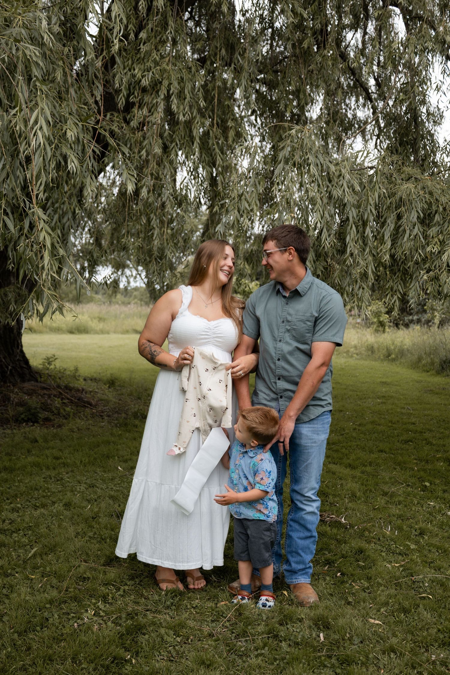 Pregnant woman in white dress laughing with husband and toddler son under a weeping willow tree.