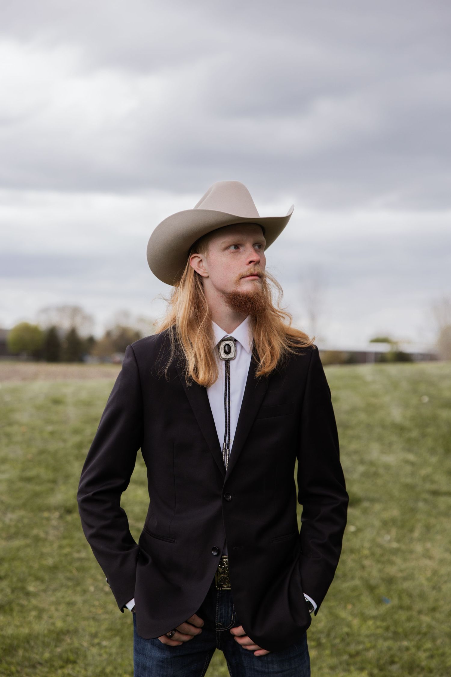 Handsome groom in black suit, bolo tie, and tan cowboy hat standing in green field under cloudy sky.