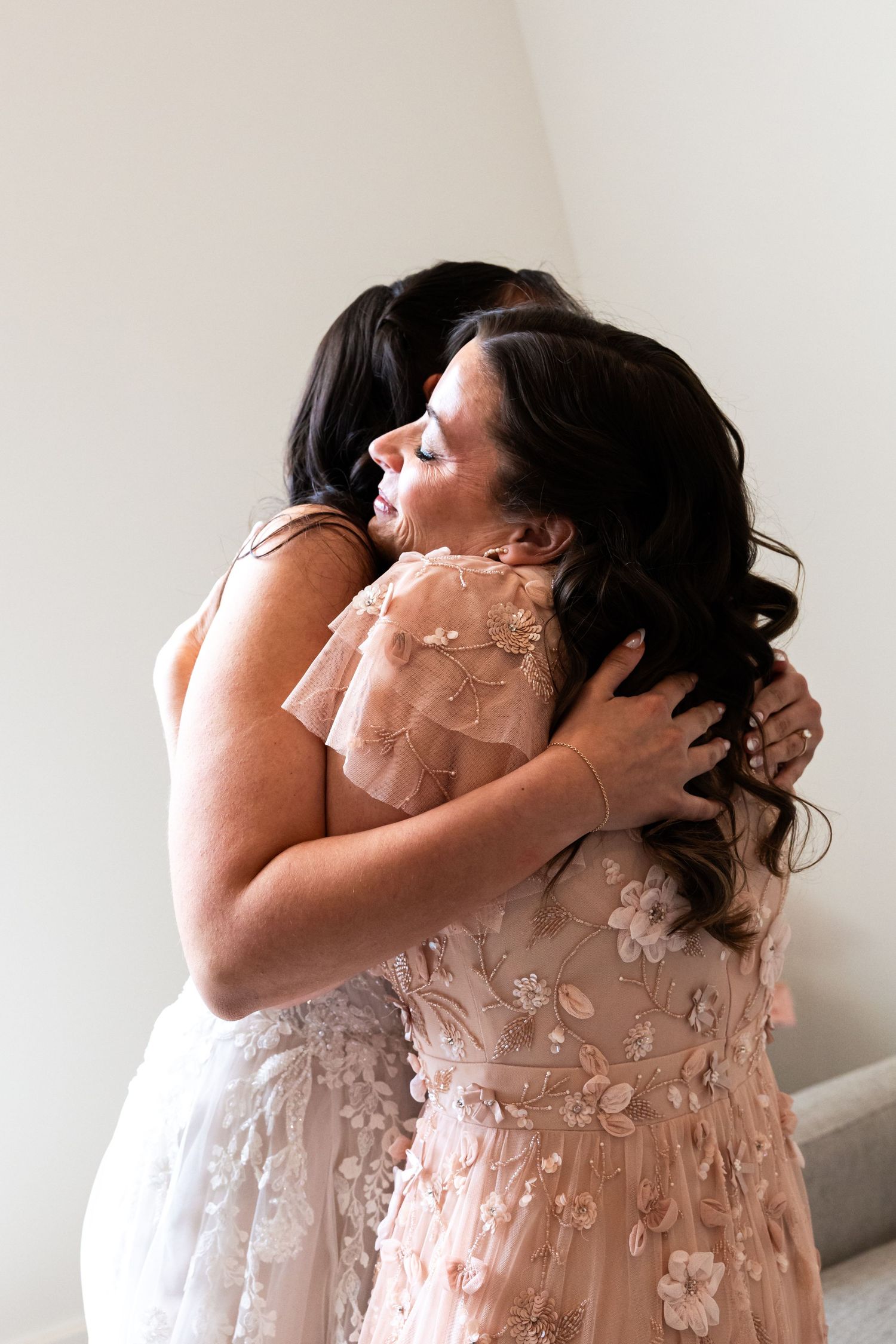 Two women share a heartfelt embrace wearing elegant floral dresses during wedding day preparations.