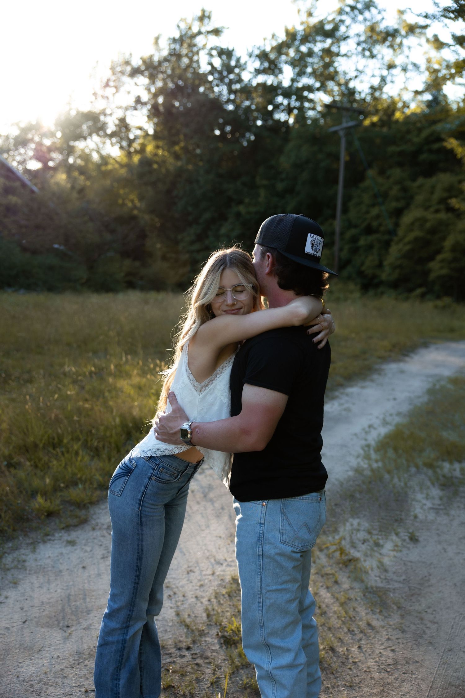 Couple embraces warmly on a sunlit gravel country road surrounded by lush green trees.