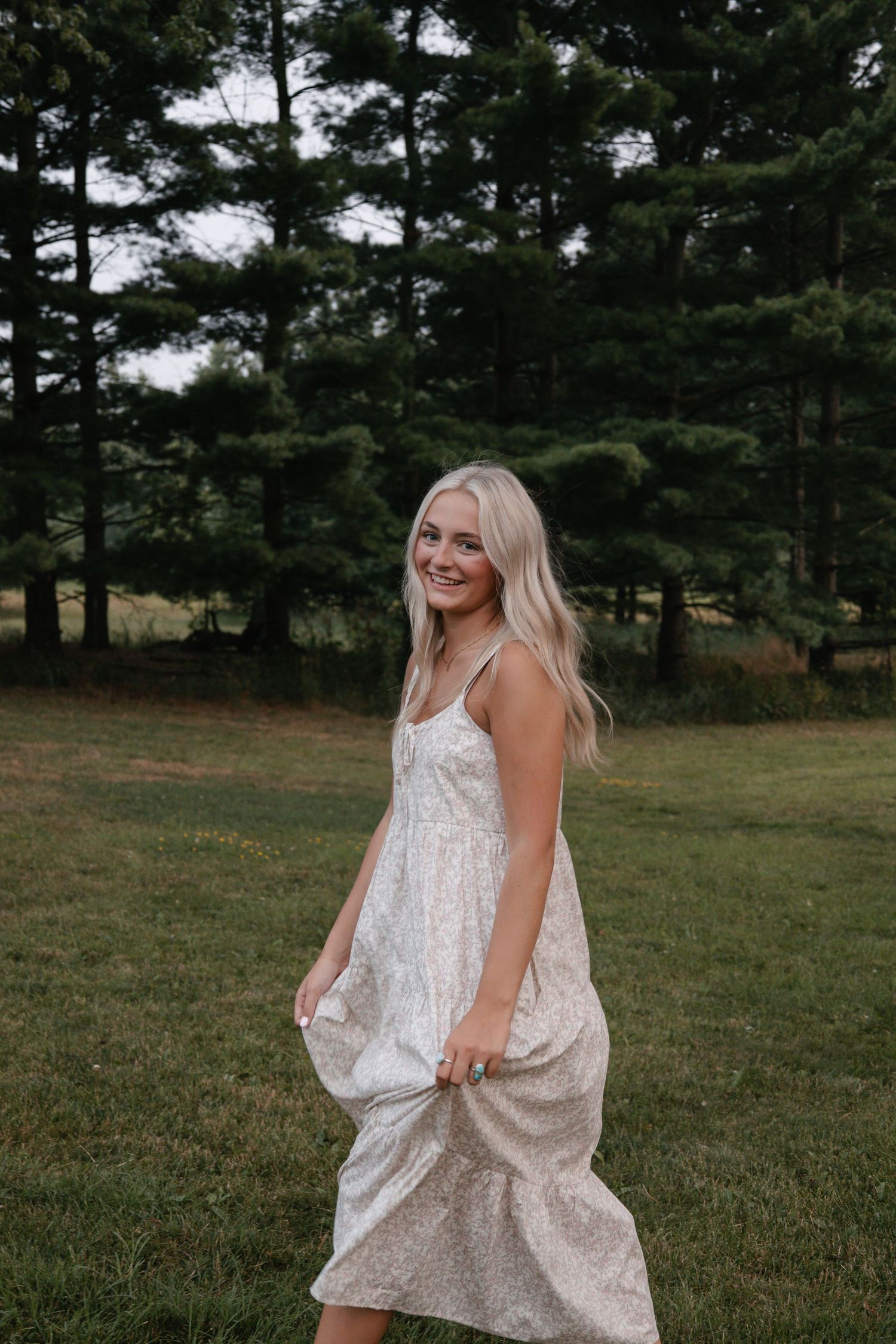 Young blonde woman in a floral sundress twirling in a green field with pine trees in the background.