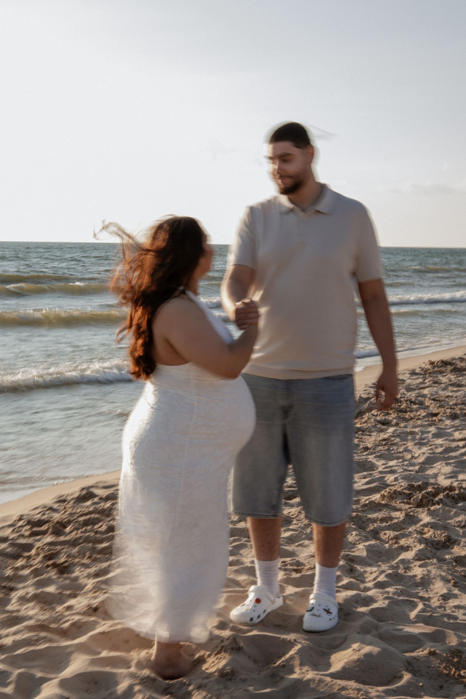A couple stands together on a sandy beach at sunset, woman in white dress, man in beige polo and denim shorts.