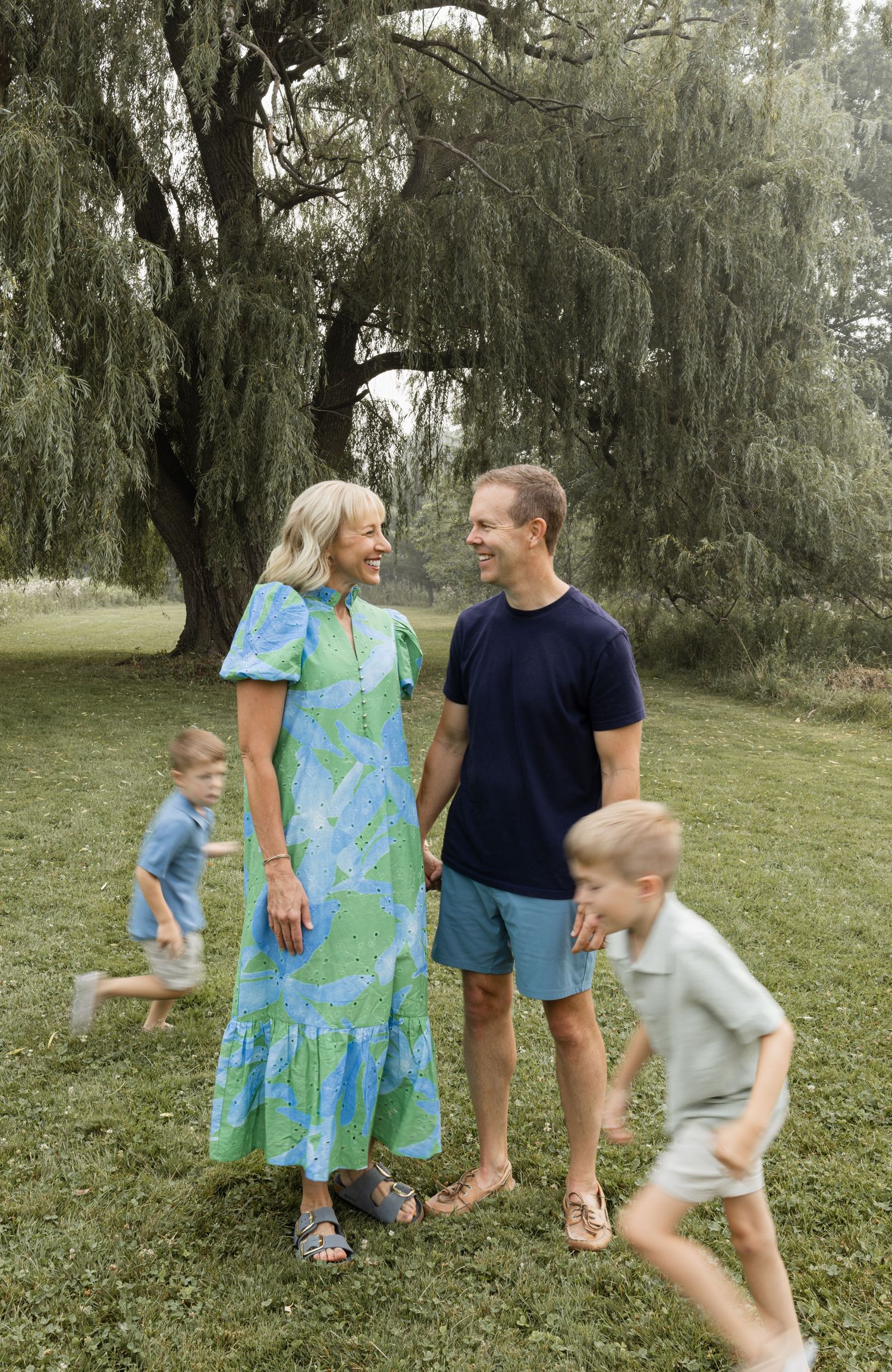 A smiling couple in a green dress and navy tee stands outdoors while two young boys run around them near a willow tree.