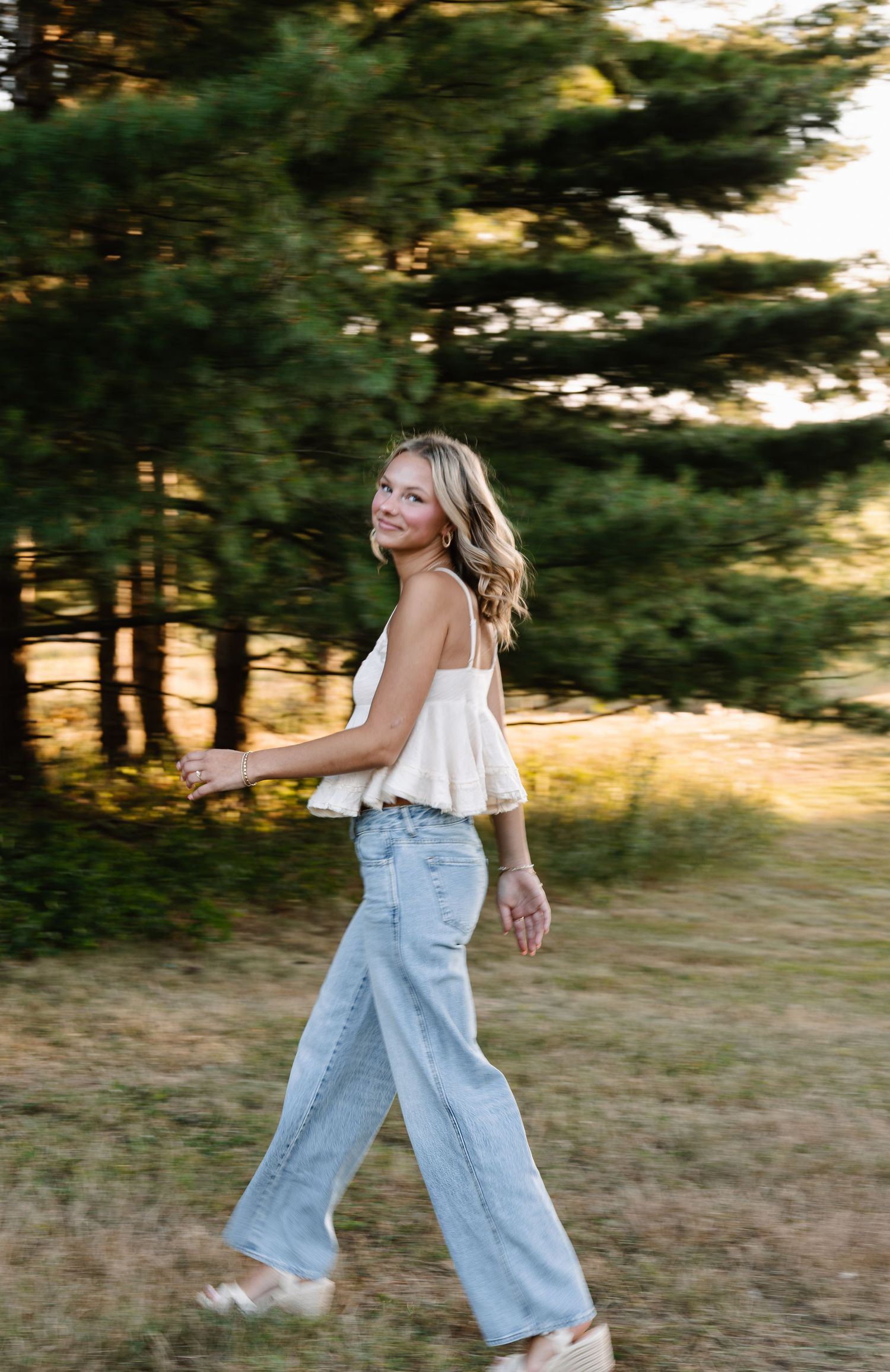 Young woman in white cami top and wide-leg jeans walking outdoors near pine trees in golden hour light.