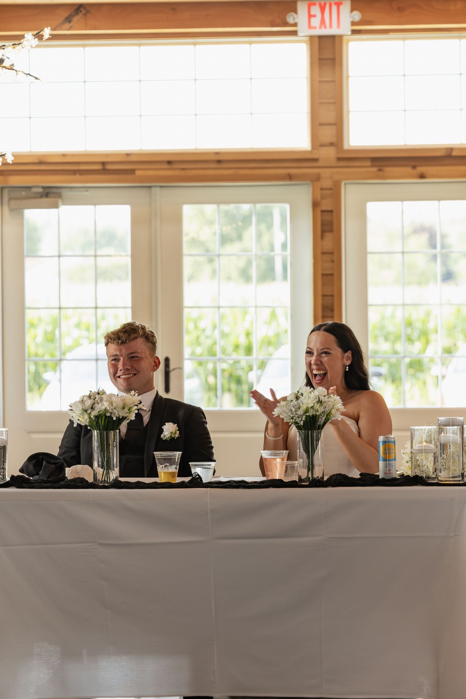 Bride and groom laughing at sweetheart table during wedding reception in bright venue.