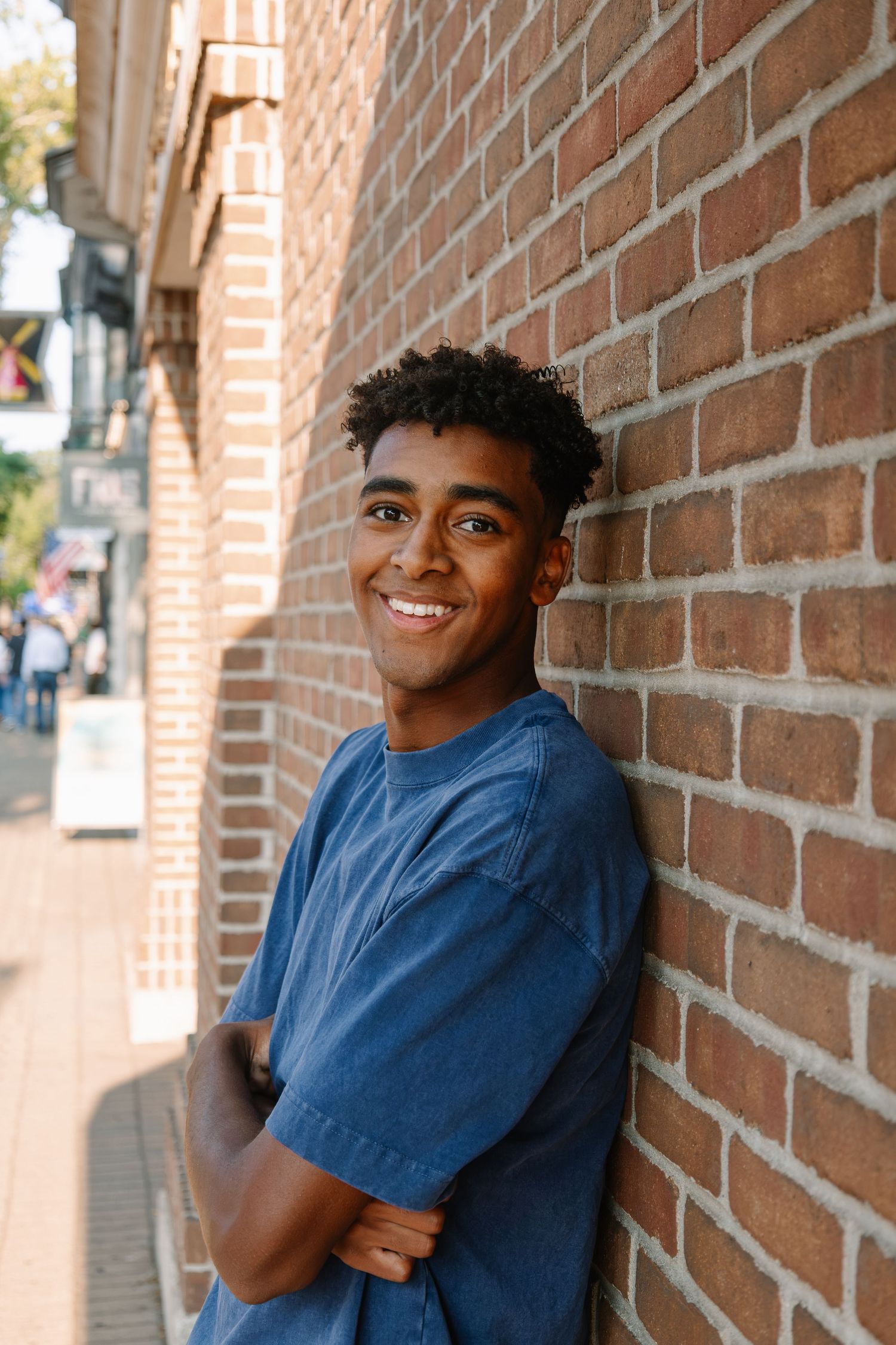 Smiling young man in blue t-shirt leaning against brick wall with arms crossed on urban street.