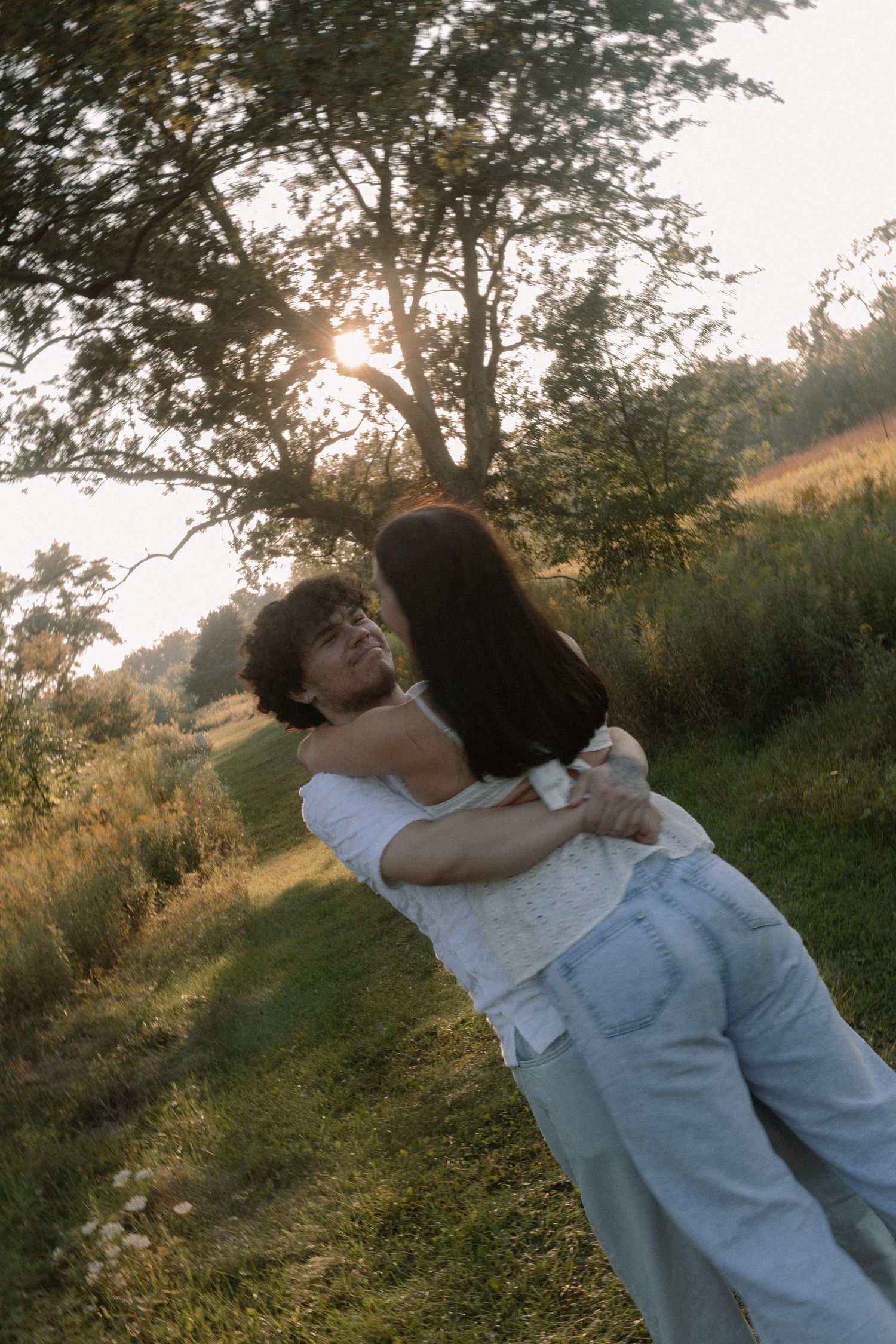 Couple embracing and spinning in a sunny green meadow with trees in the background during golden hour.