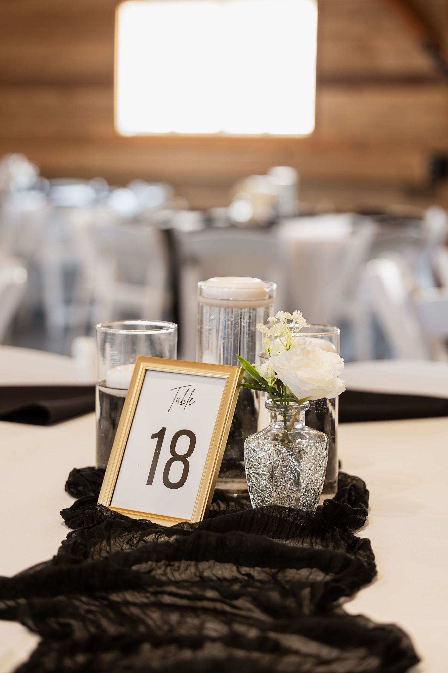 Gold-framed table number 18 centerpiece with white roses in crystal vase and candles on black lace at wedding reception.