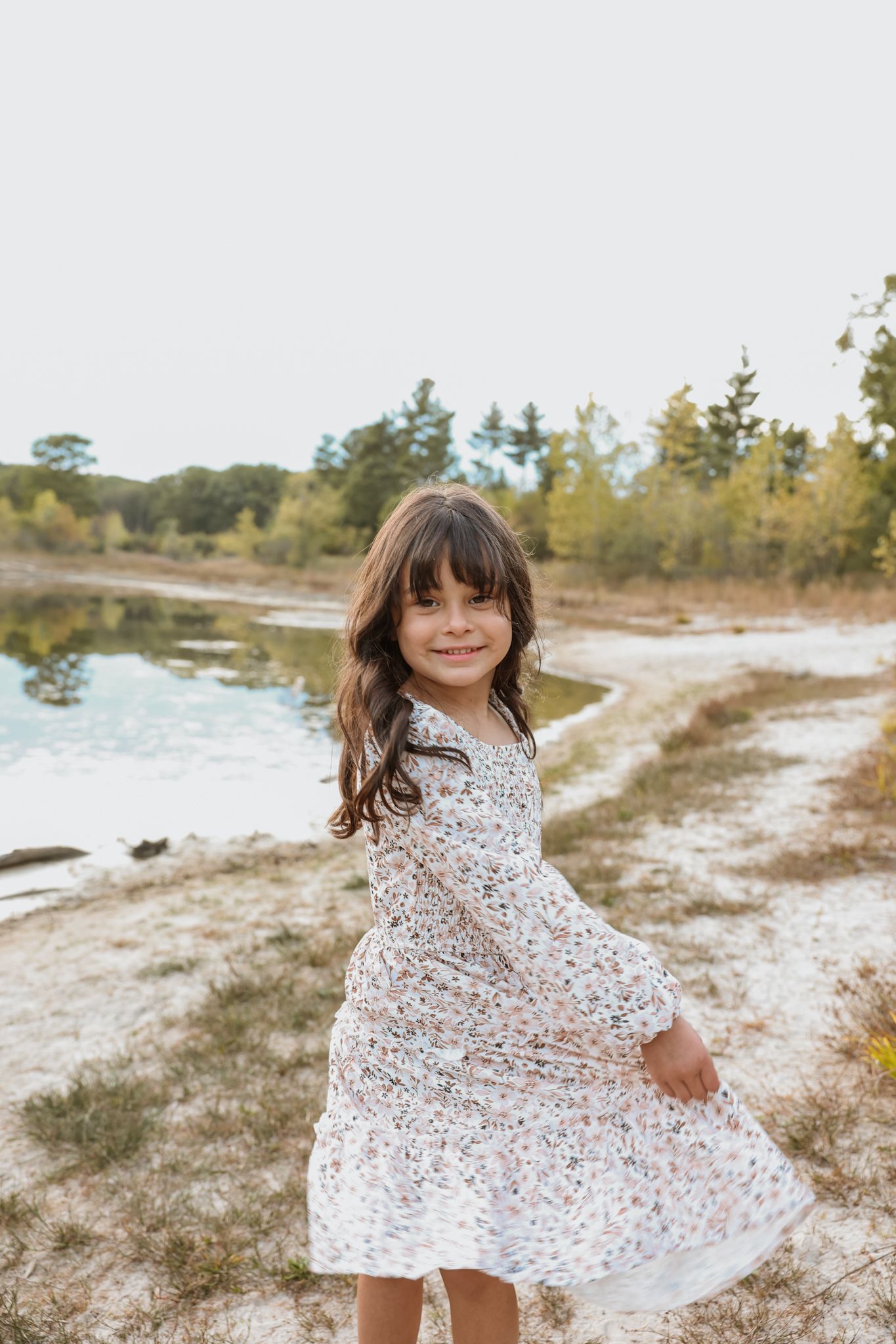 Little girl in floral dress twirls near a reflective pond surrounded by autumn trees.