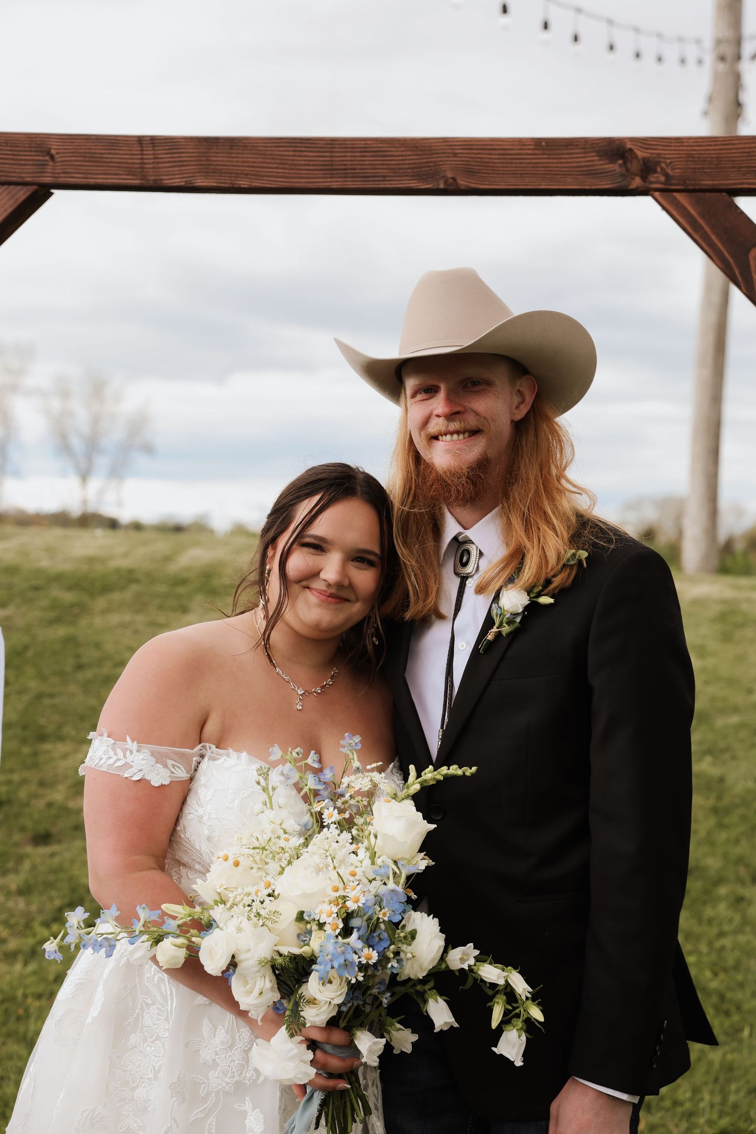Smiling newlyweds pose under wooden arch, groom in cowboy hat holding bride with blue and white bouquet.