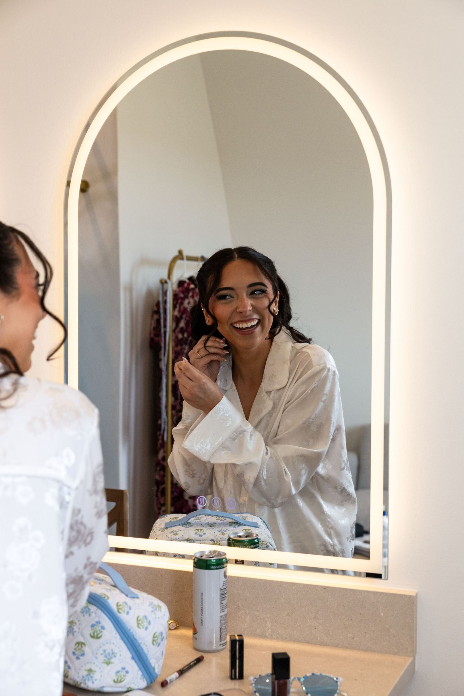 Radiant bride laughs while applying makeup at a lit vanity mirror during wedding day preparations.