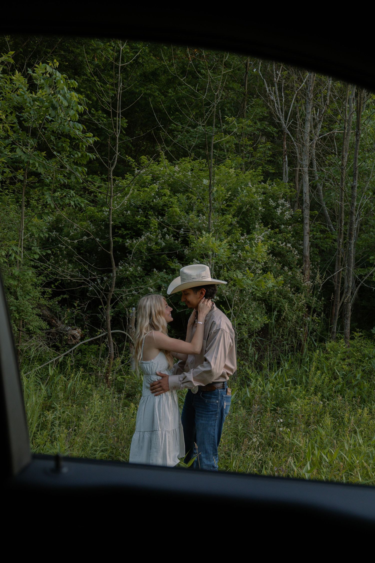 Romantic couple embracing in green forest, photographed through a car window.