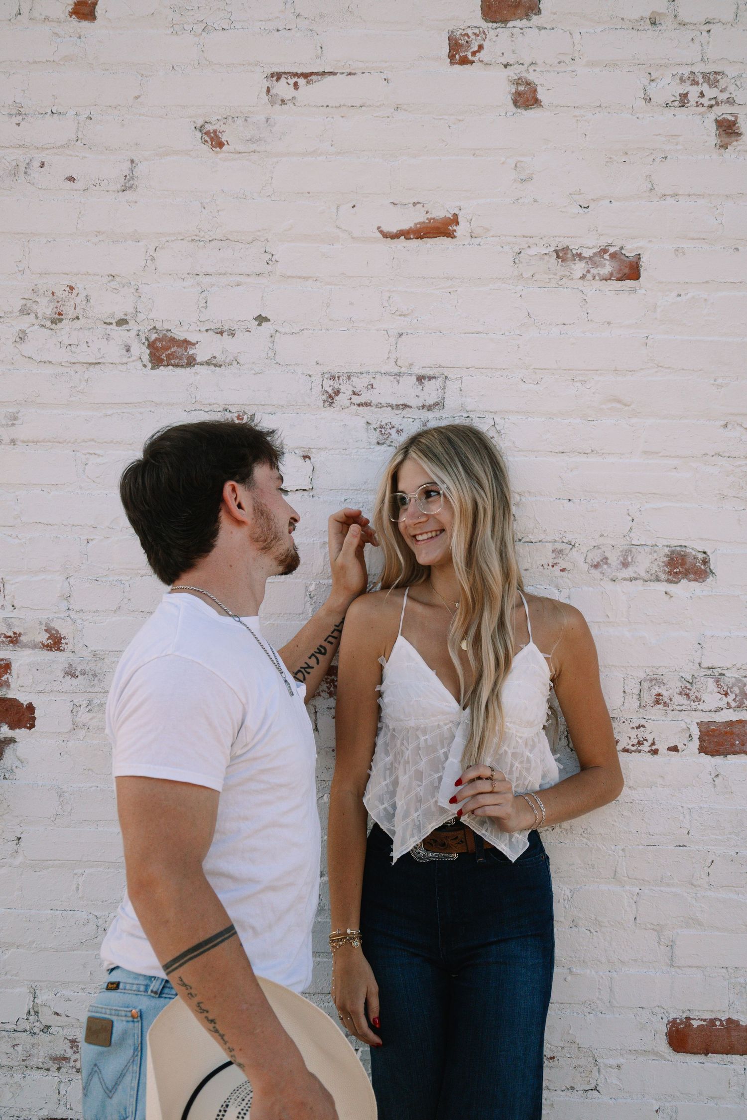 A couple smiles at each other against a white brick wall, man in white tee holding hat, woman in lace top and jeans.