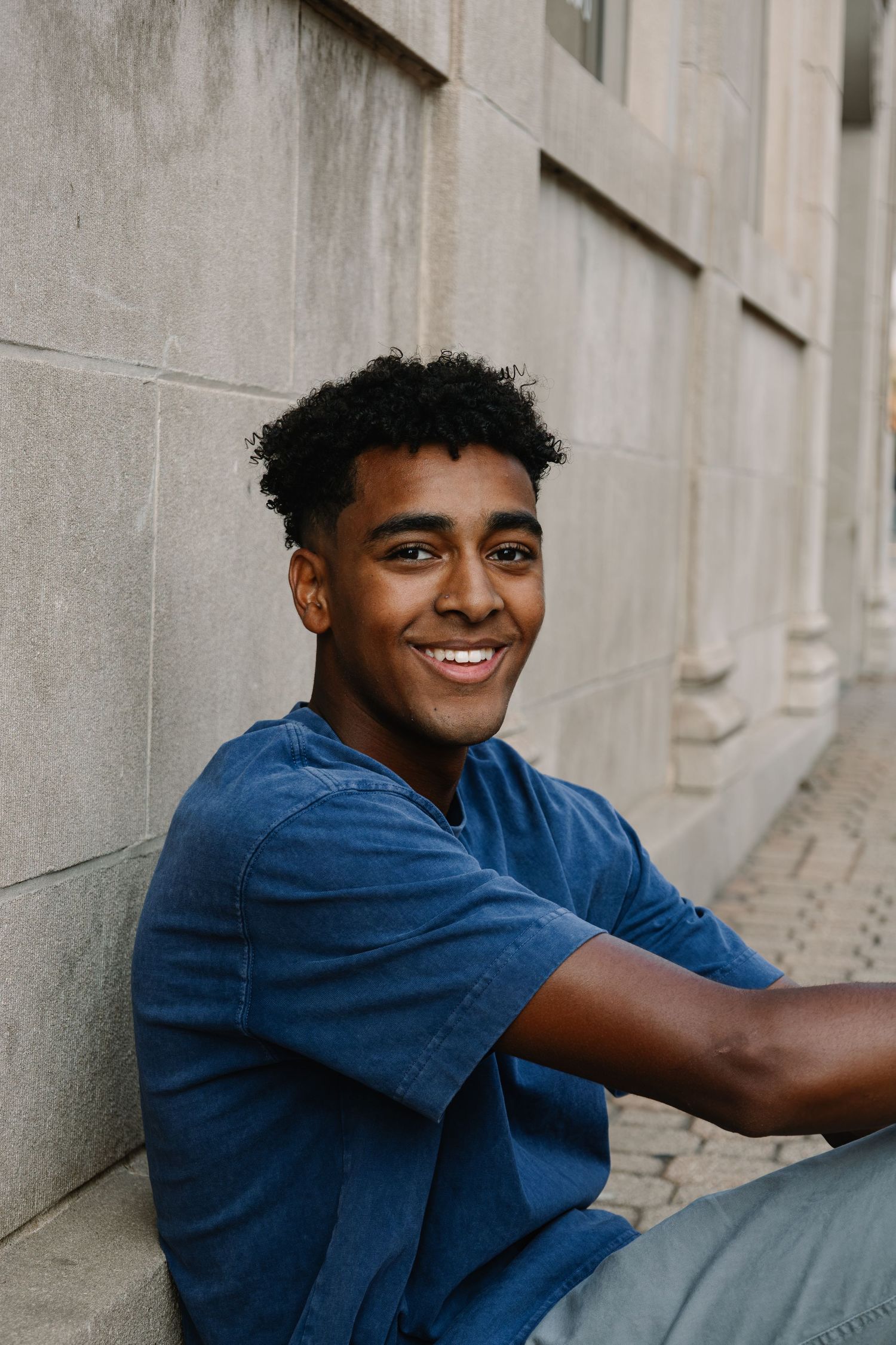 Young Black man in blue t-shirt smiling while sitting against a stone wall outdoors.