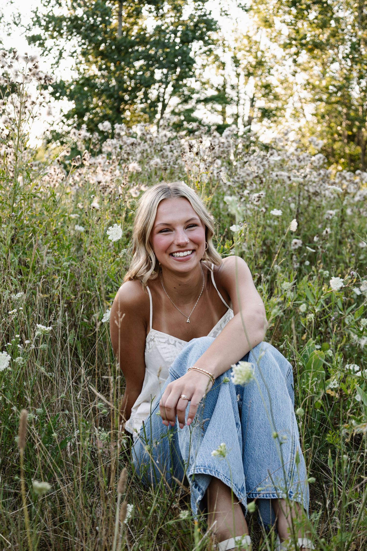 Young blonde woman smiling in a wildflower field, wearing white top and jeans, sitting among white blooms.