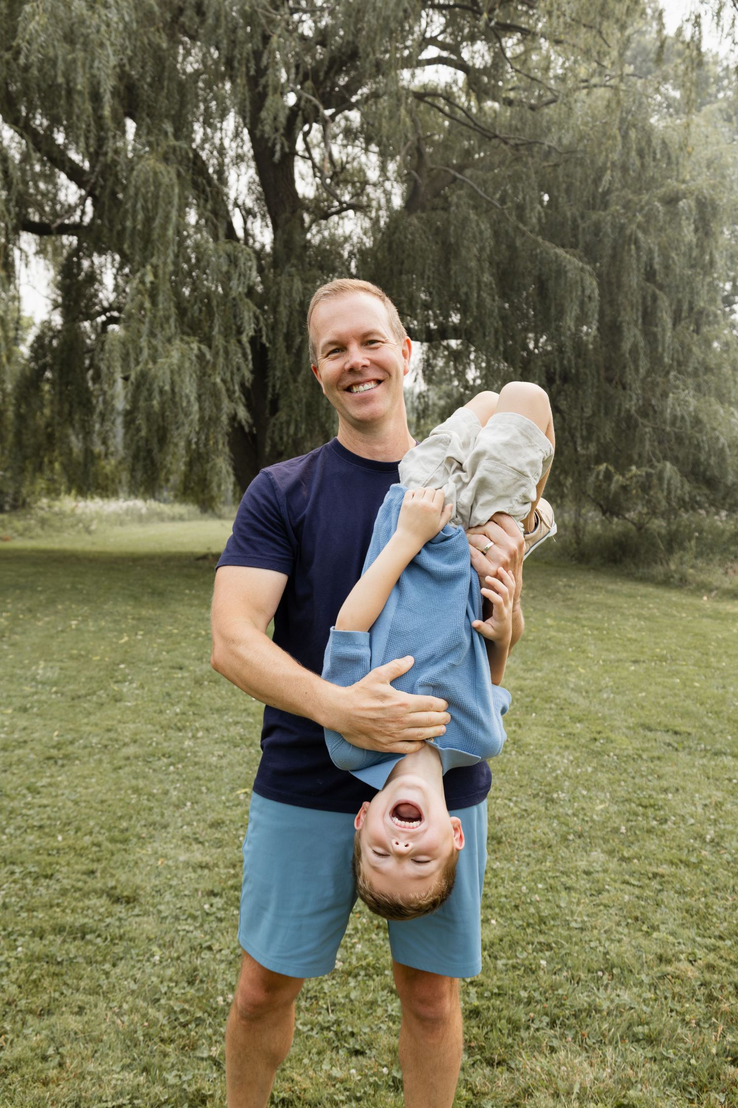 Laughing father holding young son upside down playfully in park near willow tree.