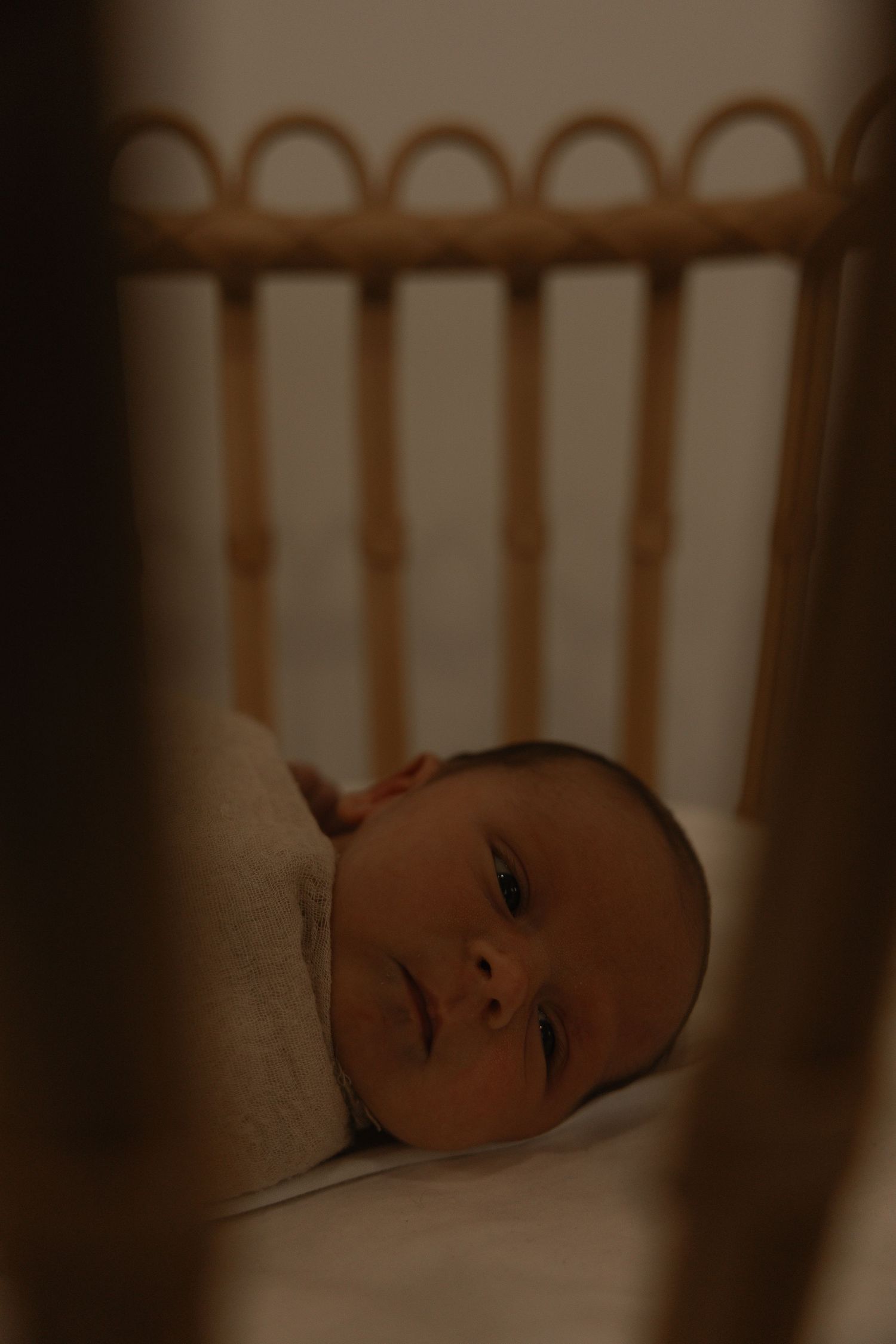Newborn baby swaddled in white blanket lying peacefully in a wooden rattan crib in dim nursery light.