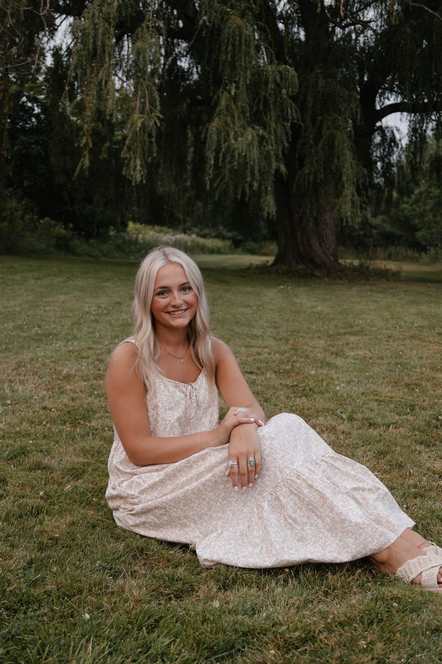 Young blonde woman in a floral midi dress sitting on green grass beneath a large weeping willow tree.