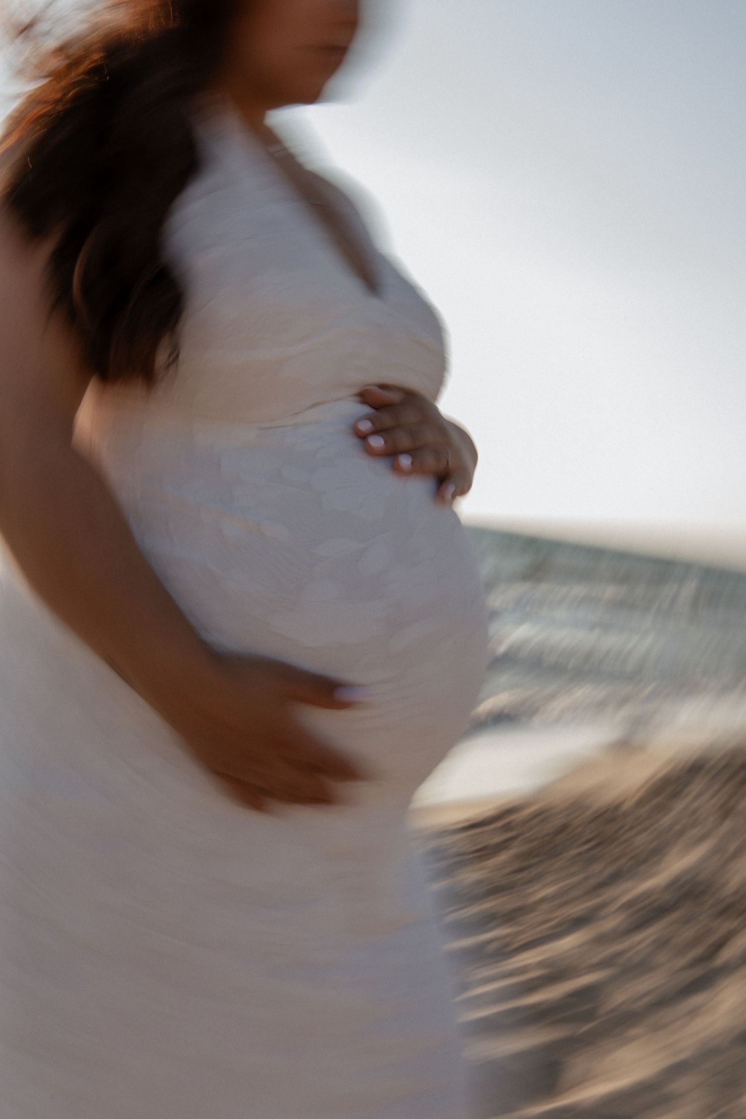 Pregnant woman in white dress cradling baby bump near ocean shore, soft blurred artistic maternity photo.