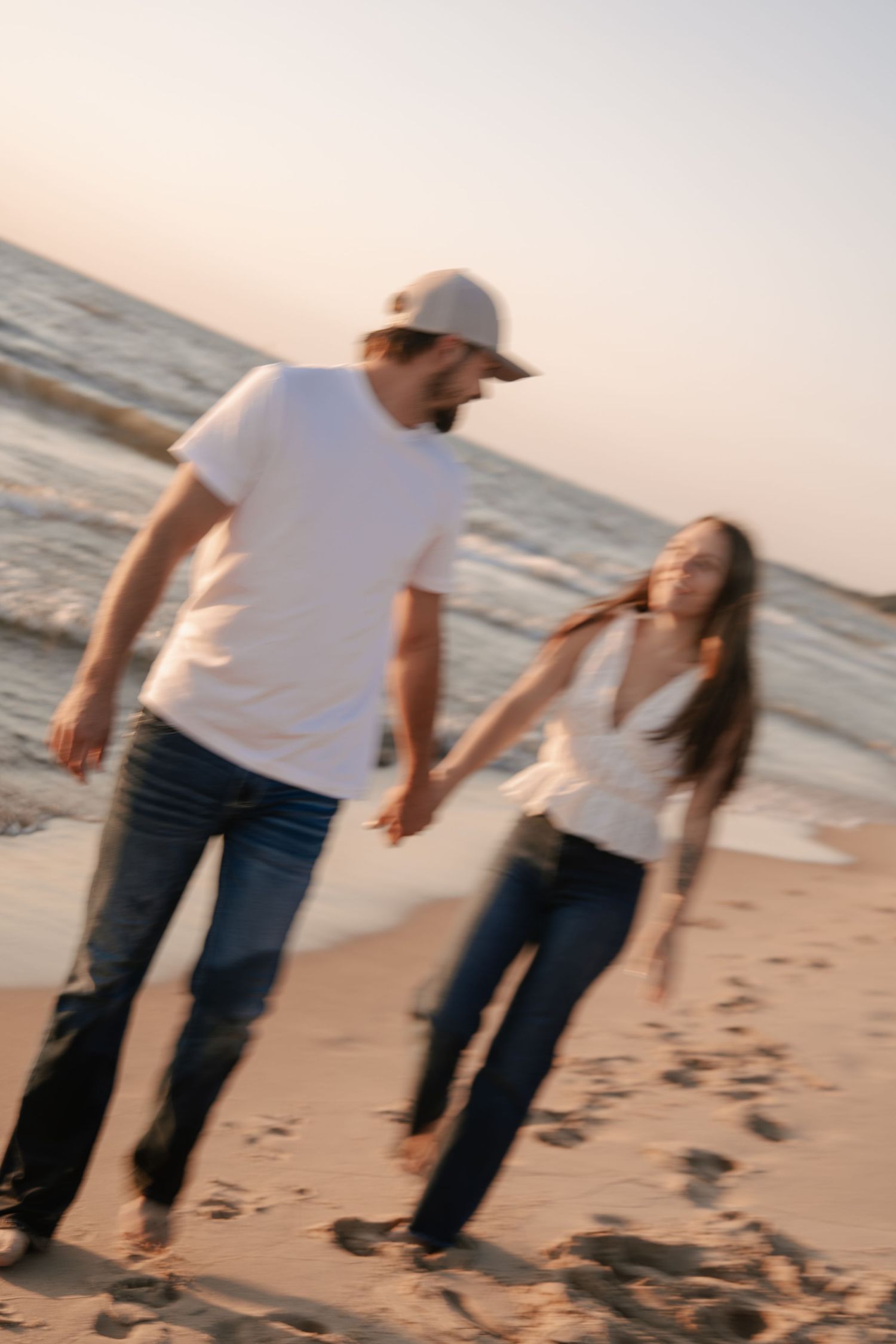 A couple holds hands walking on a sandy beach at sunset, both wearing white tops and jeans.