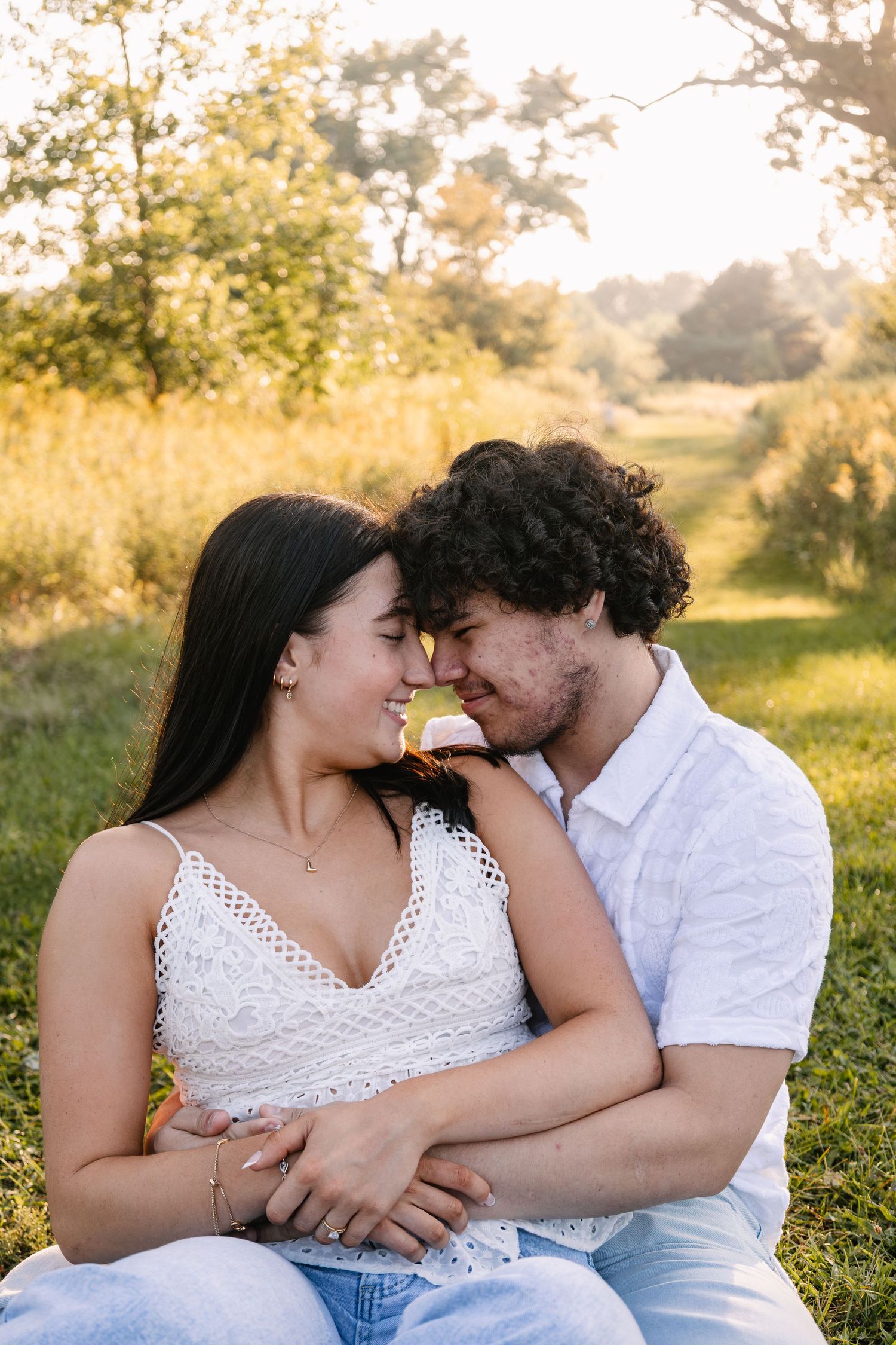 A couple sits in a sunny field, touching foreheads and embracing romantically during golden hour outdoors.