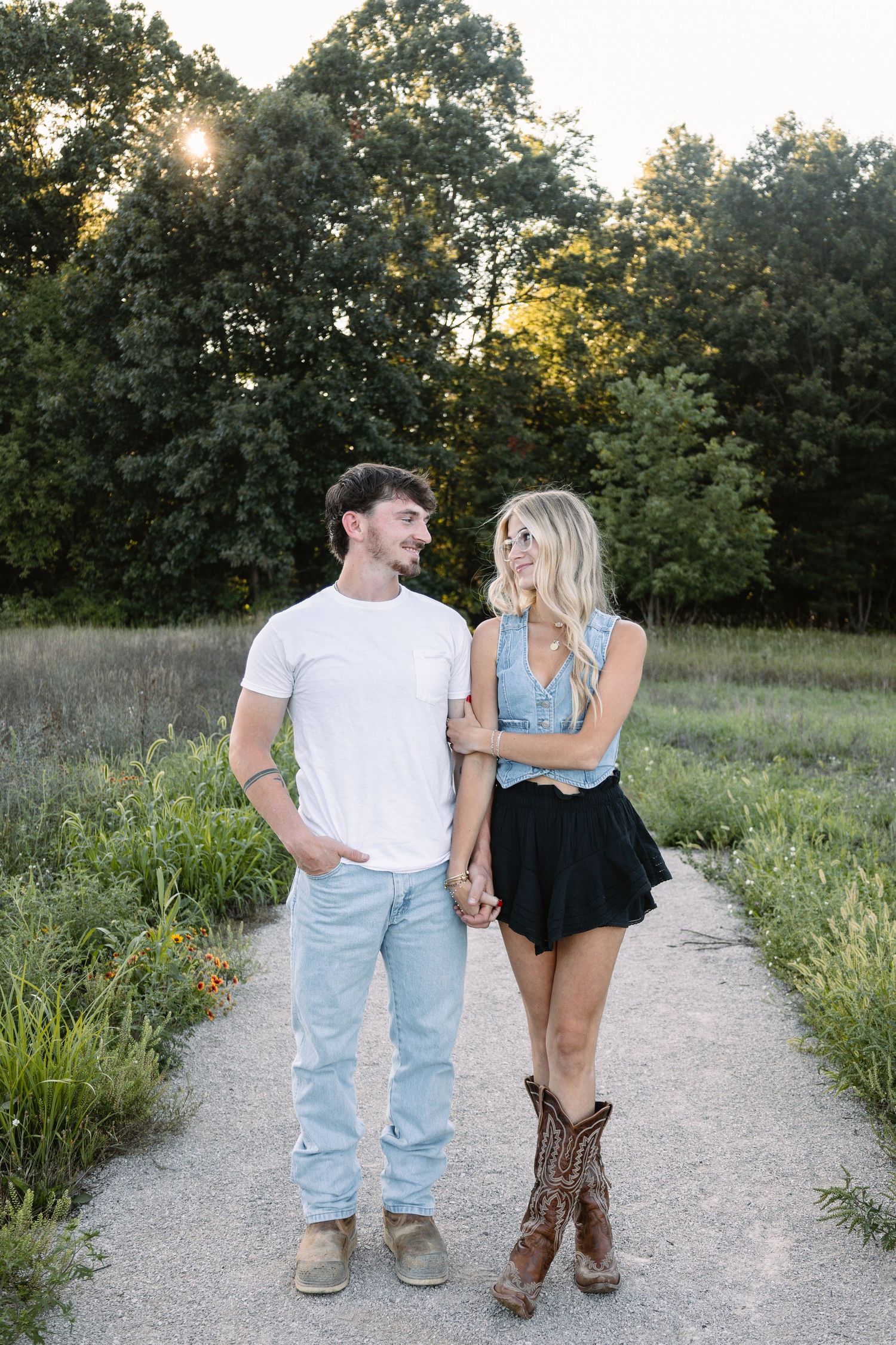 A couple holds hands on a garden path at golden hour, smiling at each other, she wears a denim top, black skirt, and cowboy boots.