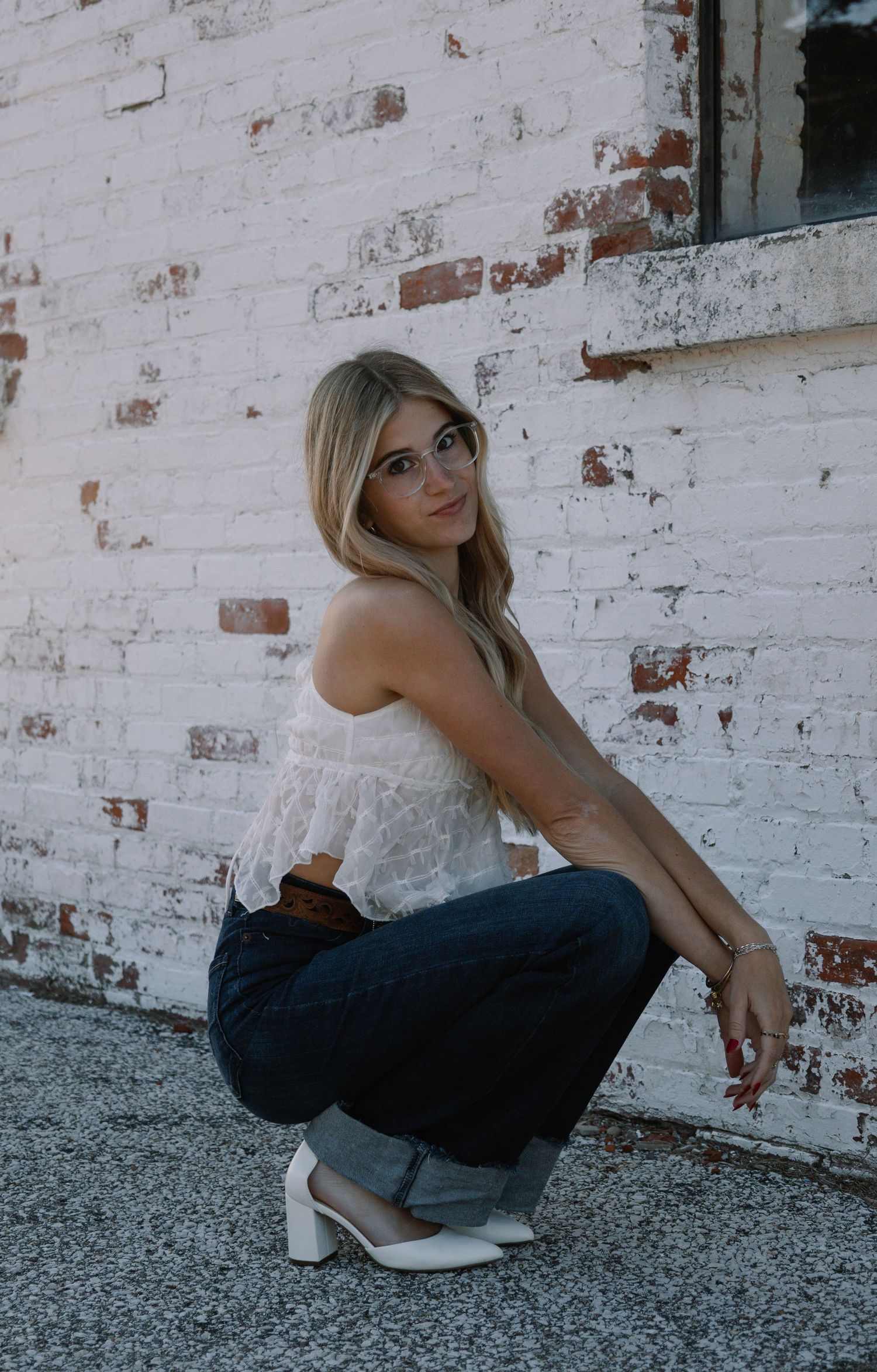Blonde woman in white lace top and dark jeans crouching by white brick wall wearing heels.