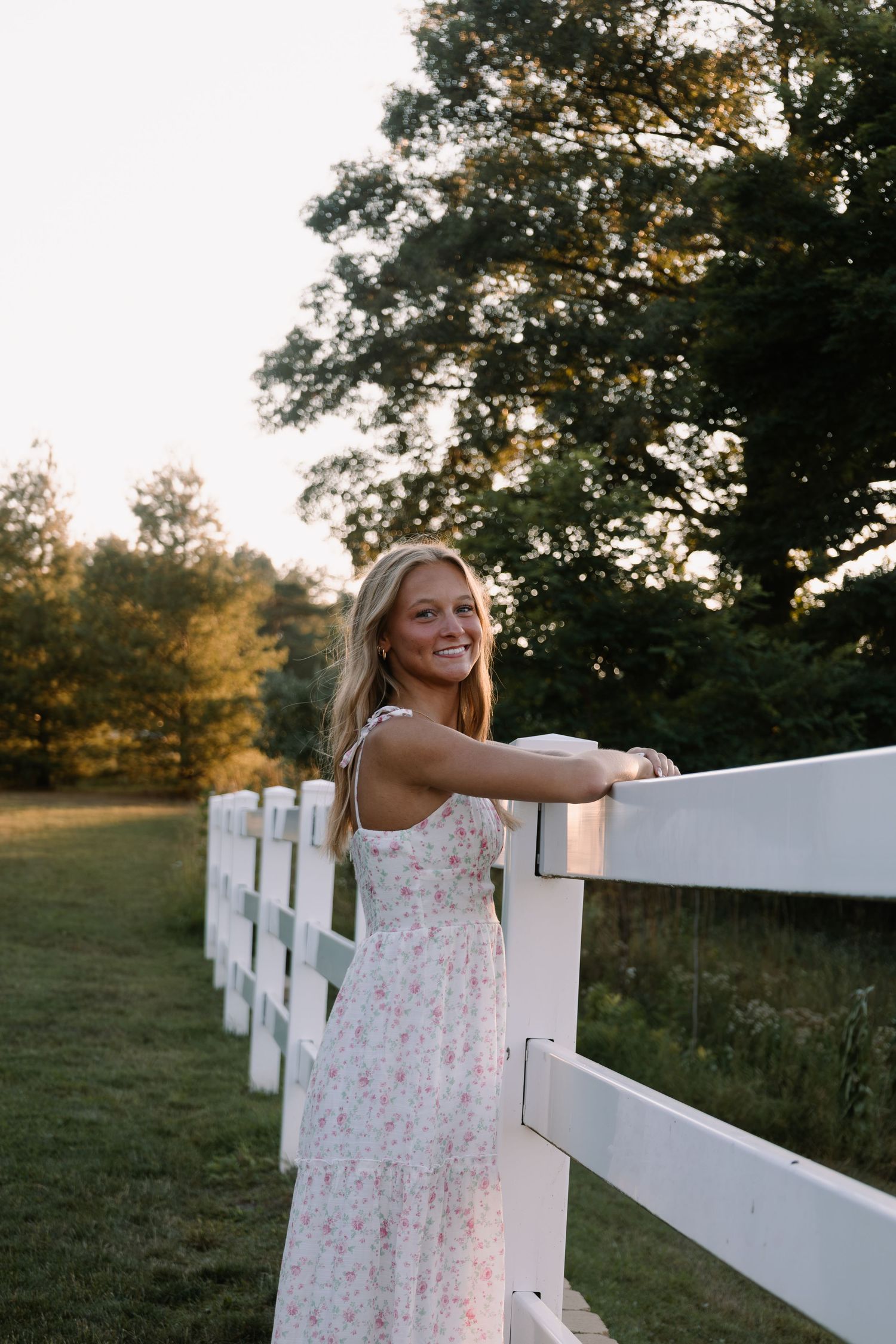 Smiling blonde woman in floral sundress leaning on white fence at golden hour with lush green trees behind her.