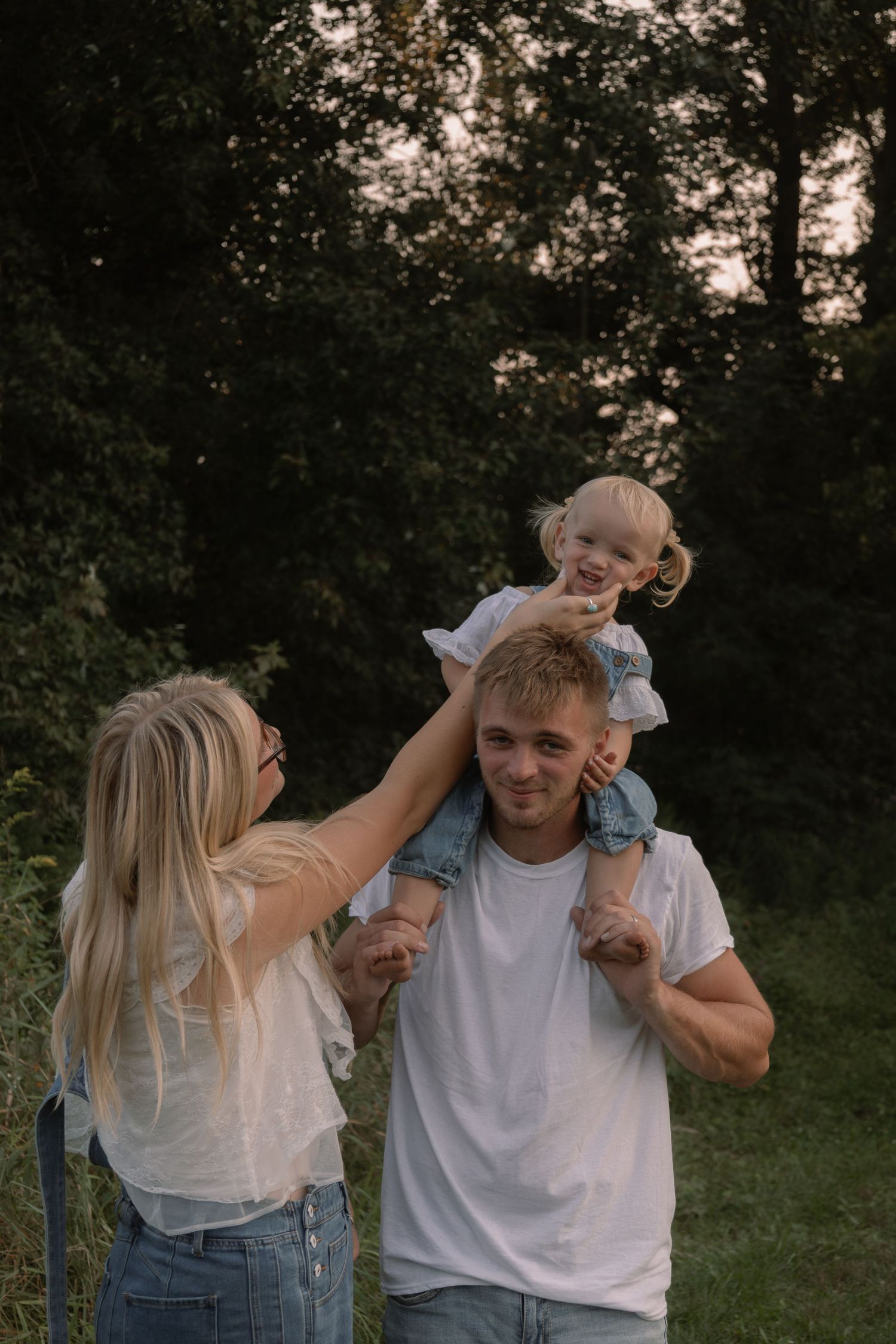 Young family enjoying outdoors, father carrying toddler on shoulders while mother smiles in green park setting.