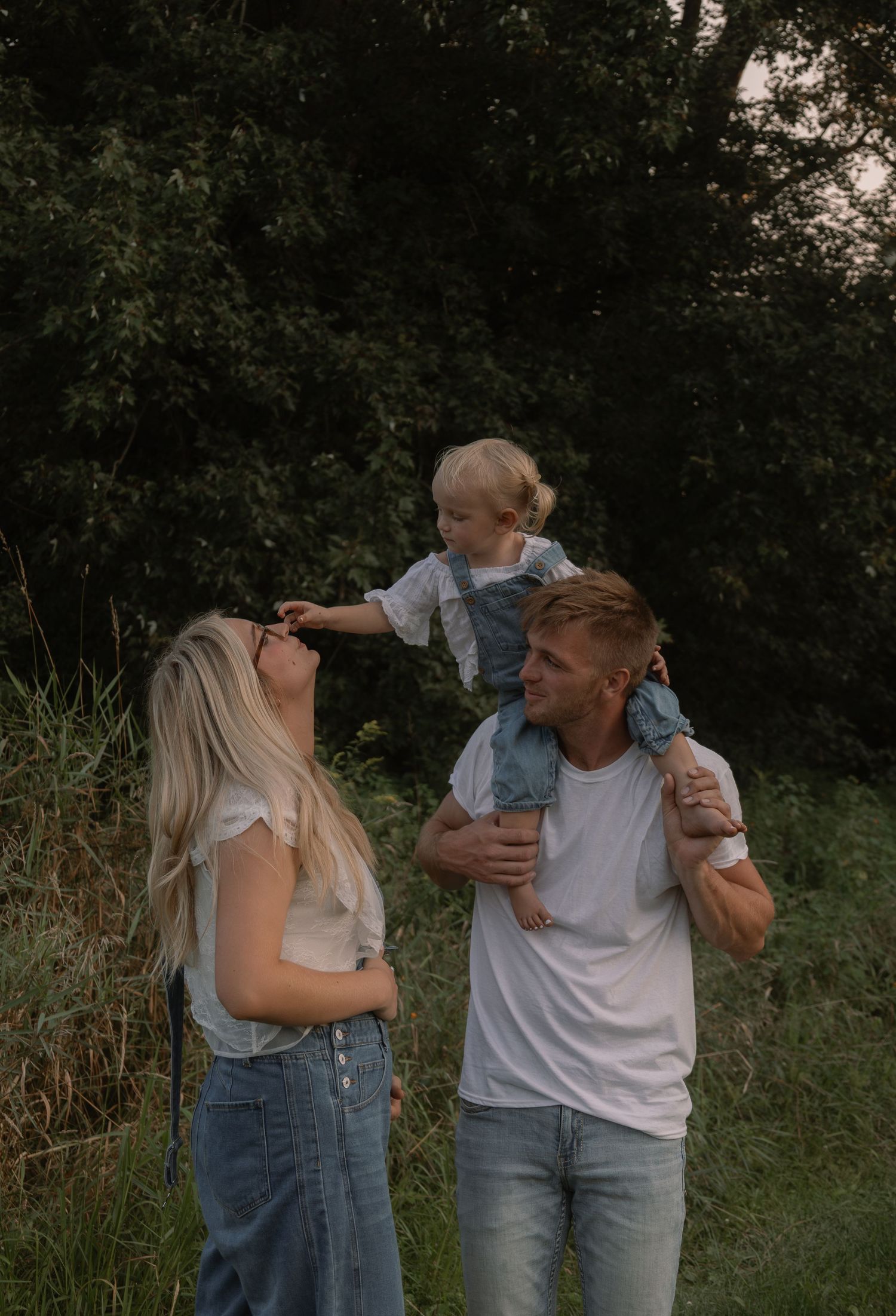 Young family with a toddler on dad's shoulders in a lush green outdoor setting at golden hour.
