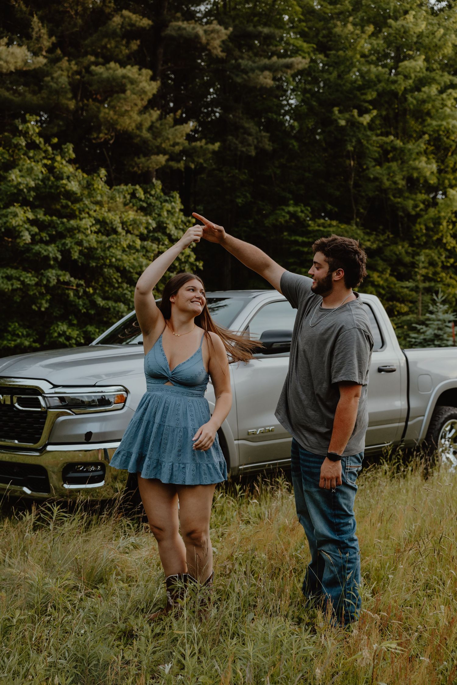 Couple dancing in a grassy field beside a silver pickup truck surrounded by lush green trees.
