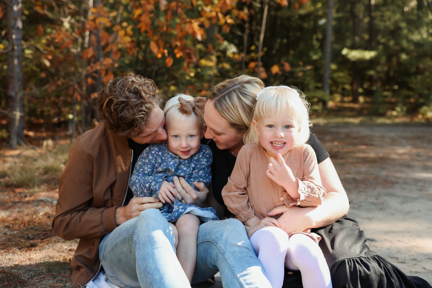 Parents lovingly kissing their two young daughters while sitting together in an autumn forest.