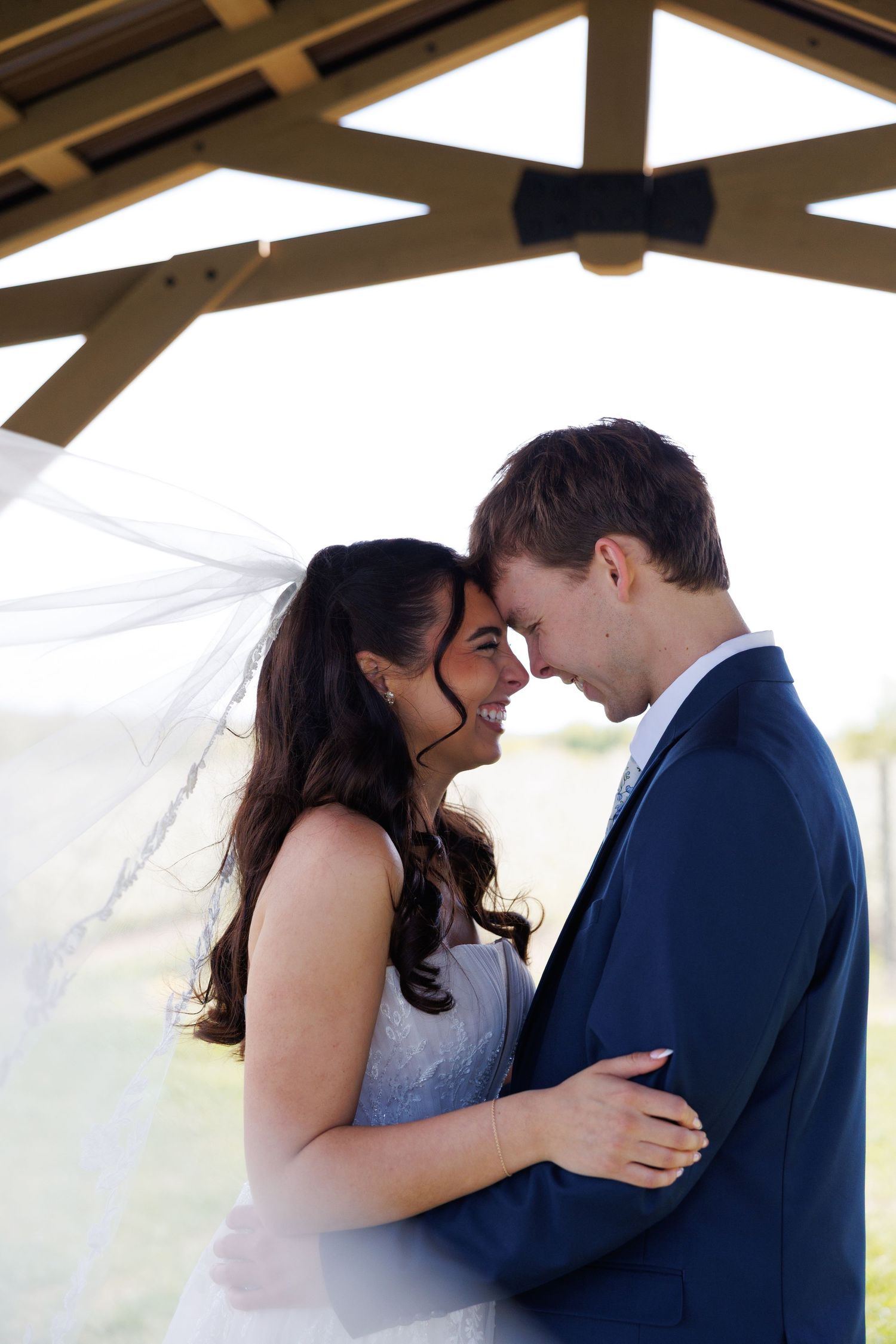 Laughing bride and groom touching foreheads under a pavilion with veil blowing in the wind.