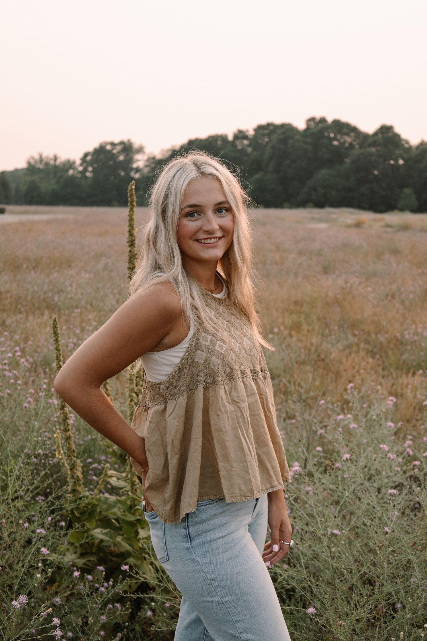 Smiling blonde woman in tan lace top and jeans posing in a wildflower meadow at golden hour.