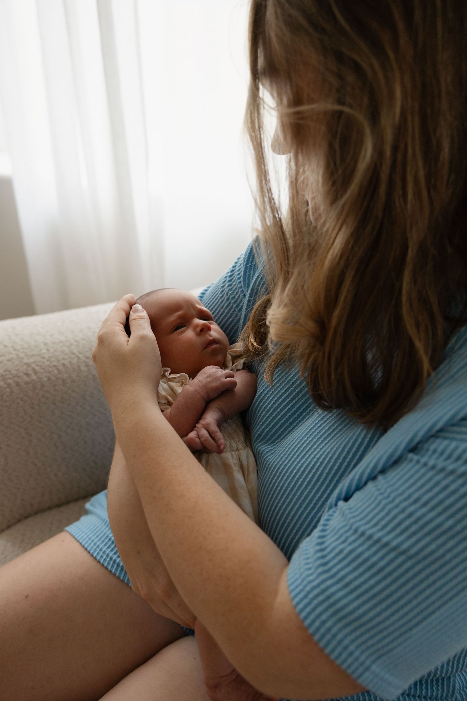 Mother in blue top cradling sleeping newborn baby while sitting in a chair near a bright window.