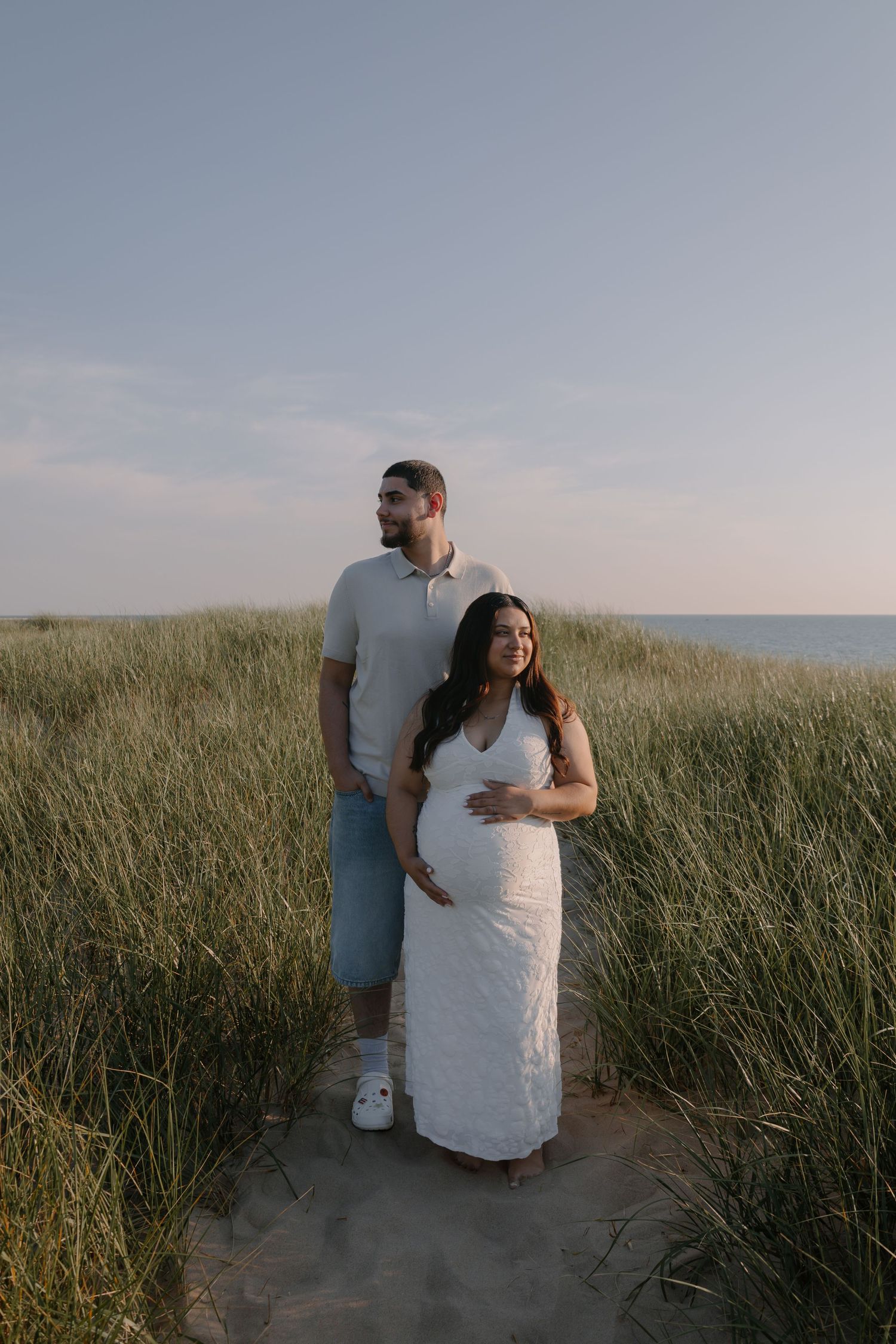 Pregnant couple standing in beach sand dunes, woman in white lace maternity dress cradling bump, golden hour light.