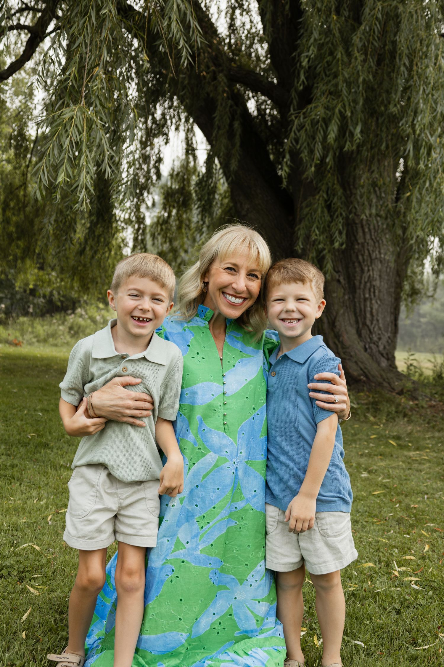 Smiling blonde mother hugging her two laughing young sons outdoors under a weeping willow tree.