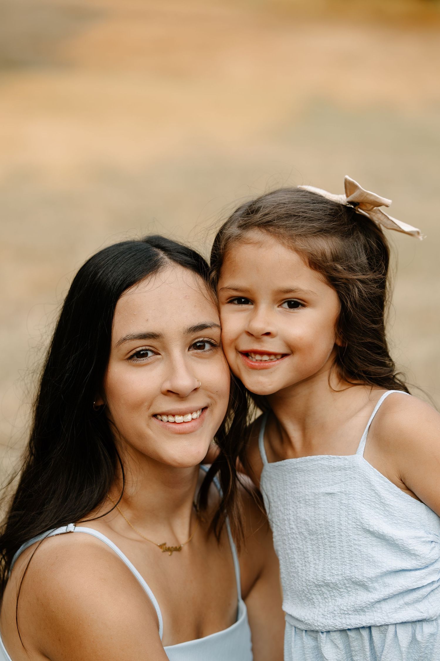 A young woman and little girl with dark hair smile together outdoors, wearing matching light blue dresses.