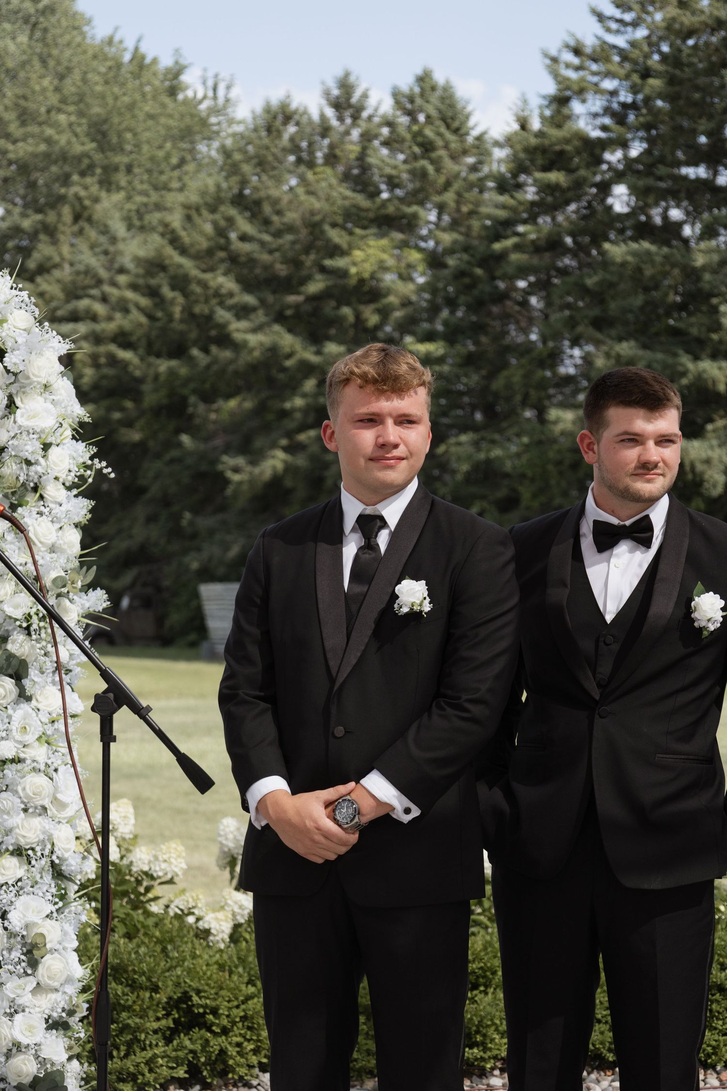 Two groomsmen in black tuxedos with white boutonnieres stand at outdoor wedding ceremony altar beside floral arch.